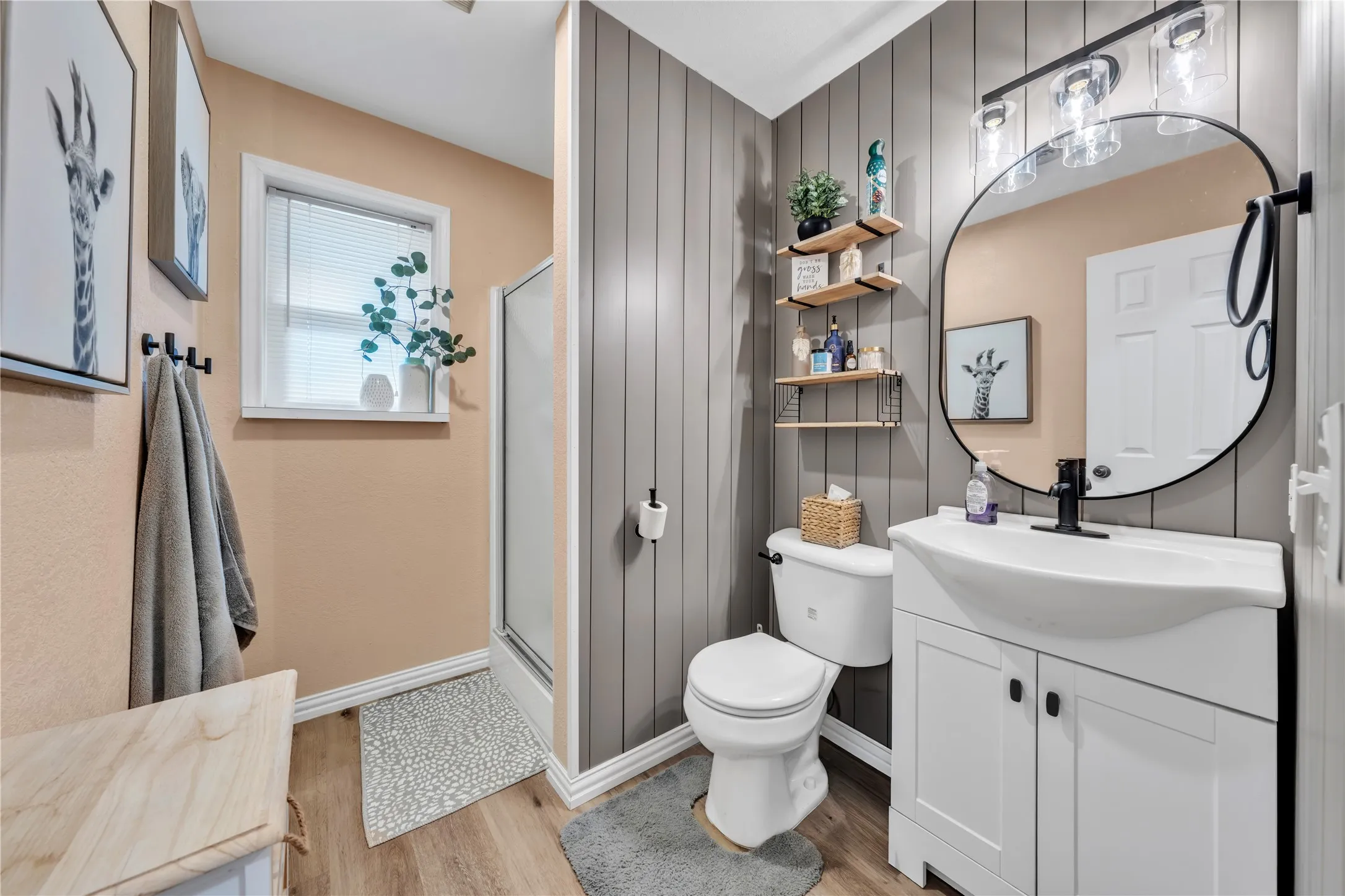 Bathroom featuring a white vanity with integrated sink, wood-finish flooring, a glass-enclosed shower, a window, and vertical panel accent wall