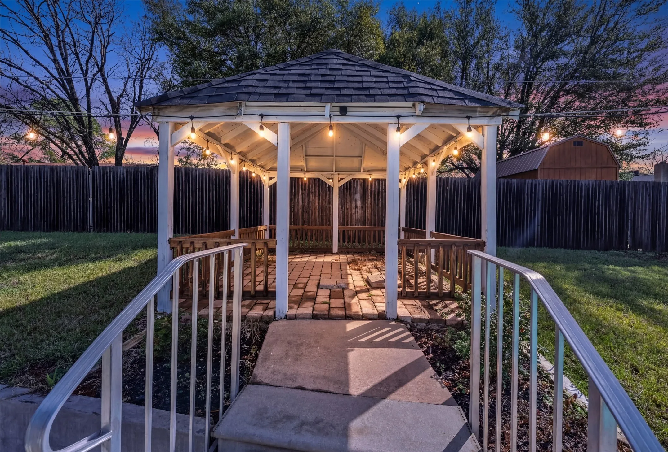 White wooden gazebo with a shingled roof and string lighting, featuring a brick-paved floor and built-in benches