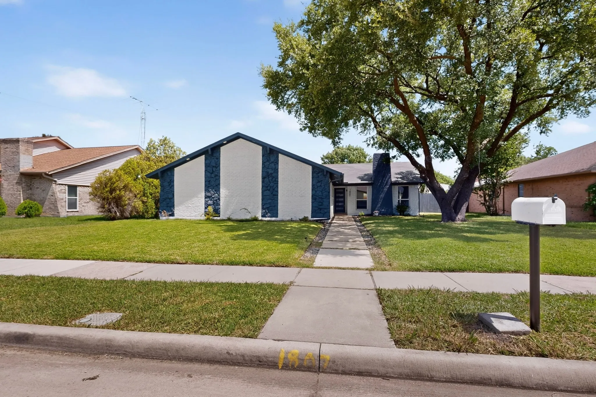 Single-story residence featuring a white and dark blue facade, a mature tree, and a concrete walkway leading to the entrance