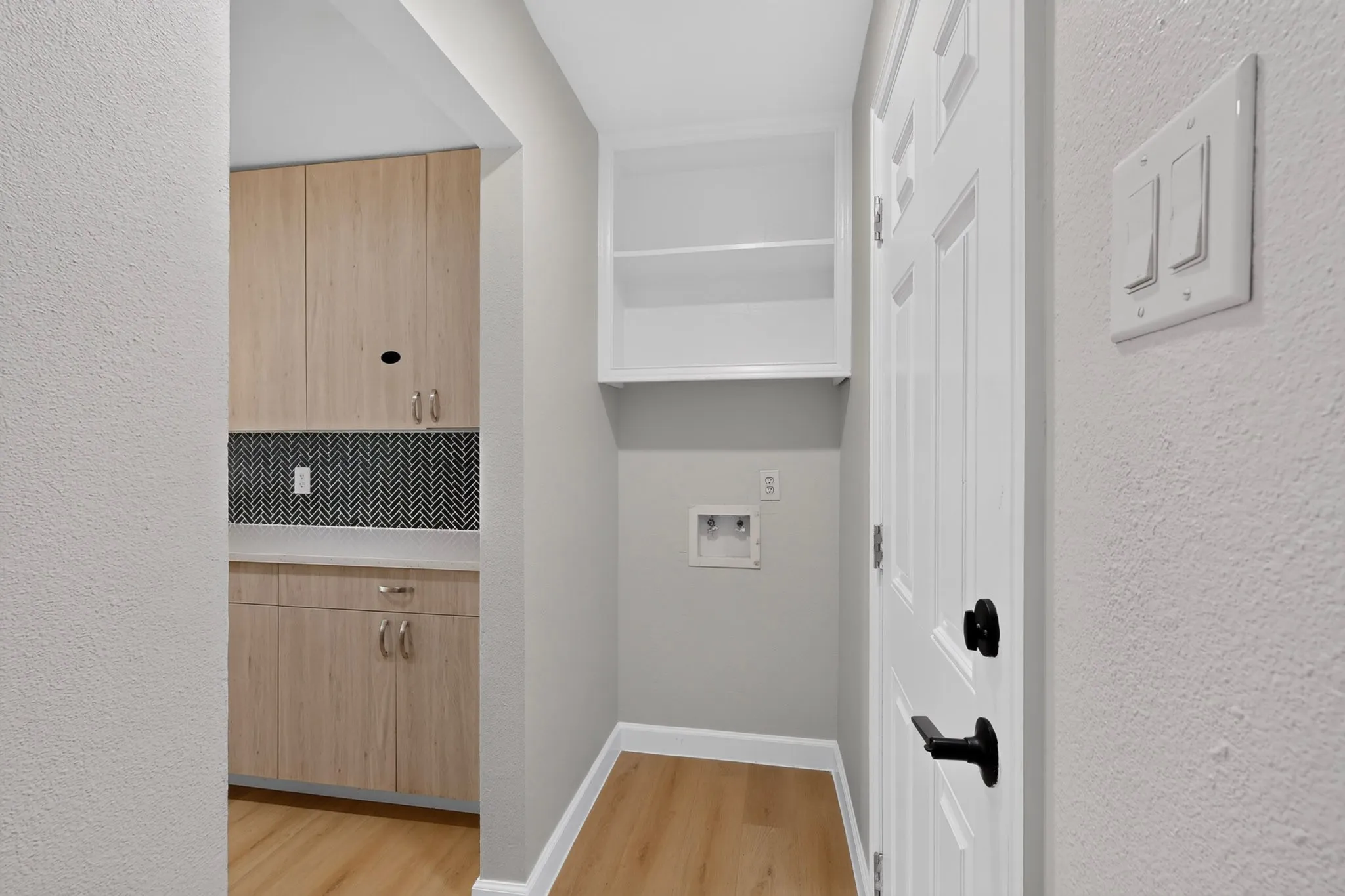 Laundry area with wood-finish flooring, light wood cabinetry, and a black and white herringbone tile backsplash