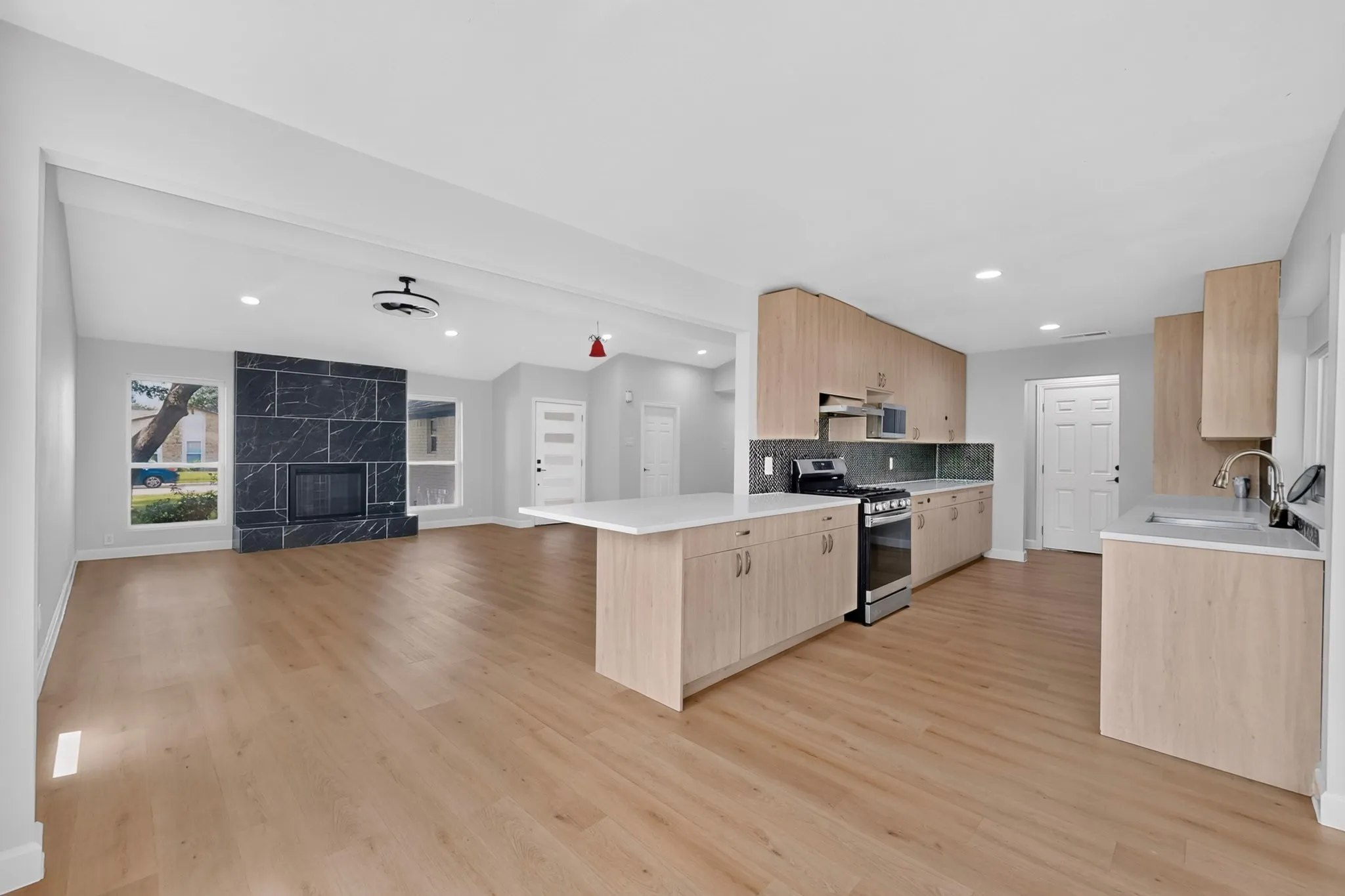 Open-concept living space featuring wood-finish flooring throughout, a modern kitchen with light wood cabinetry, white countertops, and stainless steel appliances, and a black stone-clad fireplace