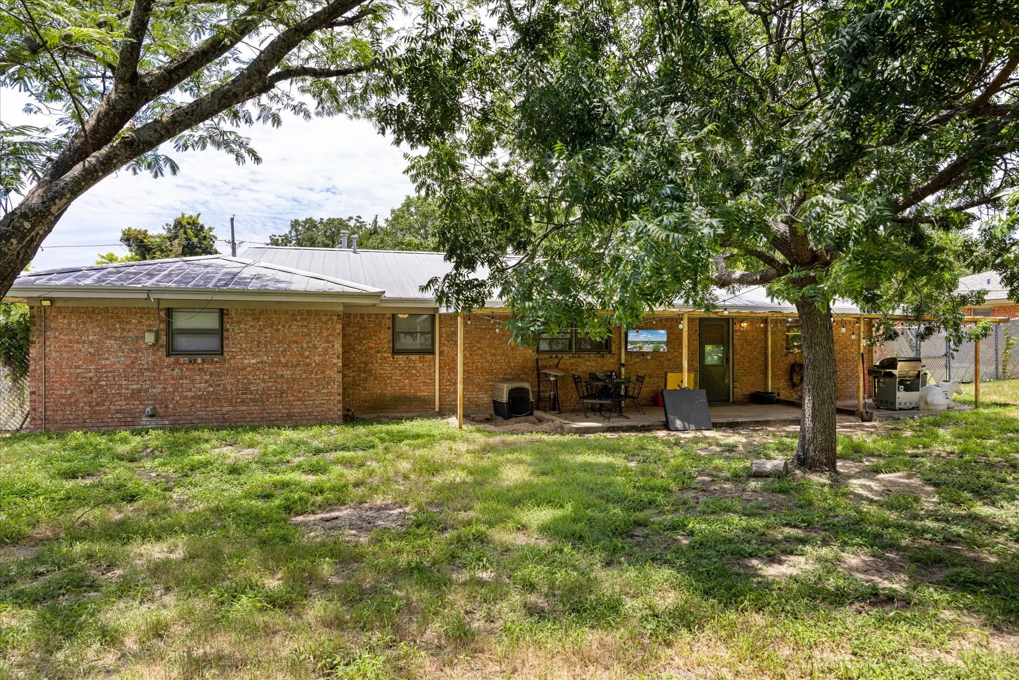 Expansive backyard featuring a mature shade tree, brick exterior, metal roof, covered patio, and chain-link fencing