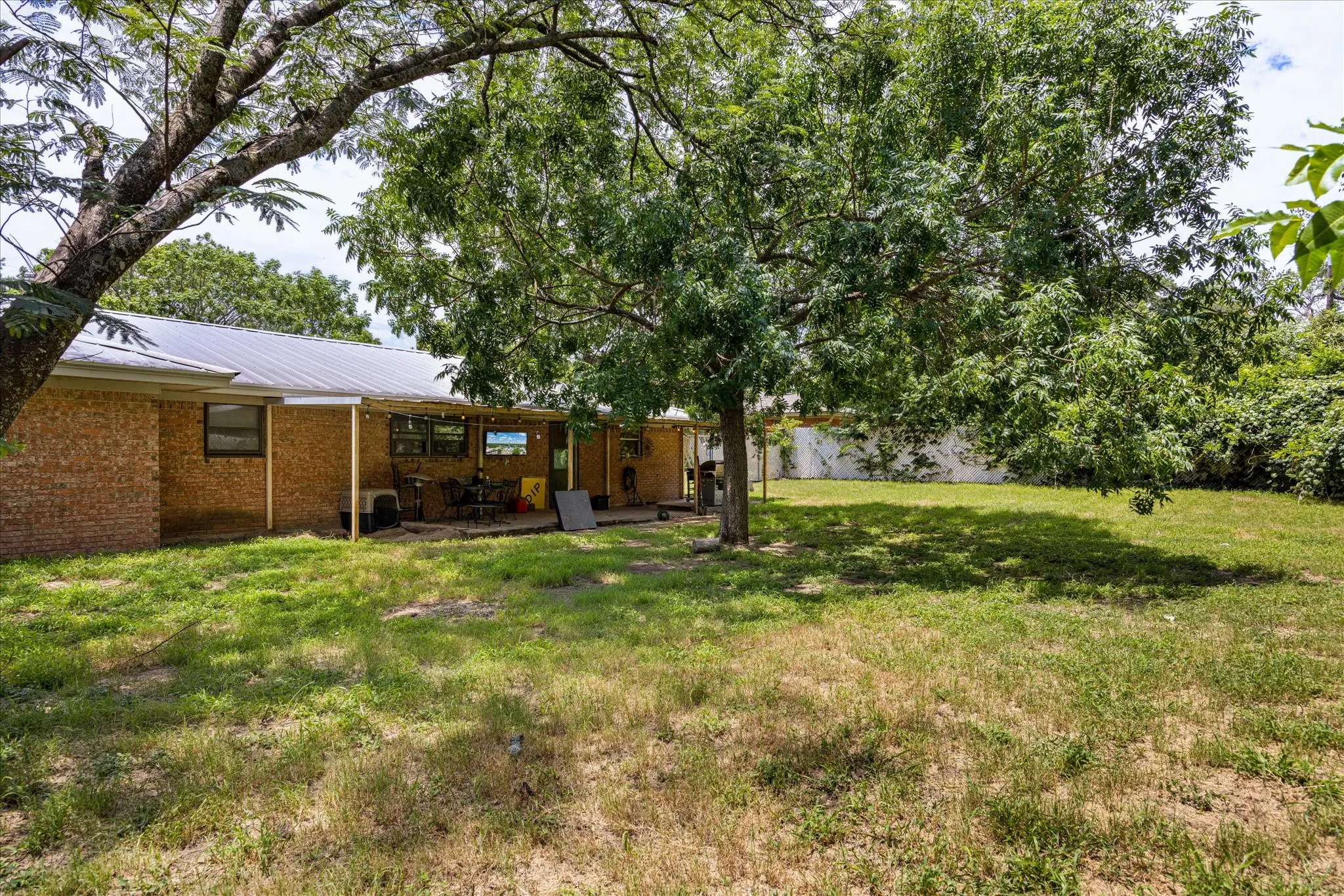 Expansive backyard featuring mature trees, a covered patio, brick exterior, and a metal roof
