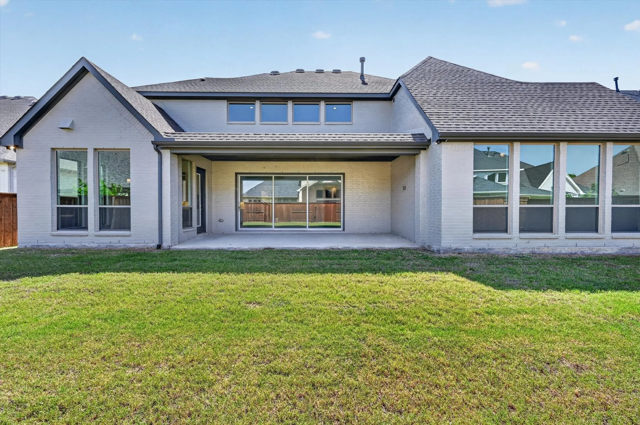 Spacious rear yard featuring a covered patio, extensive lawn, white brick exterior, multiple large windows, and a sliding glass door