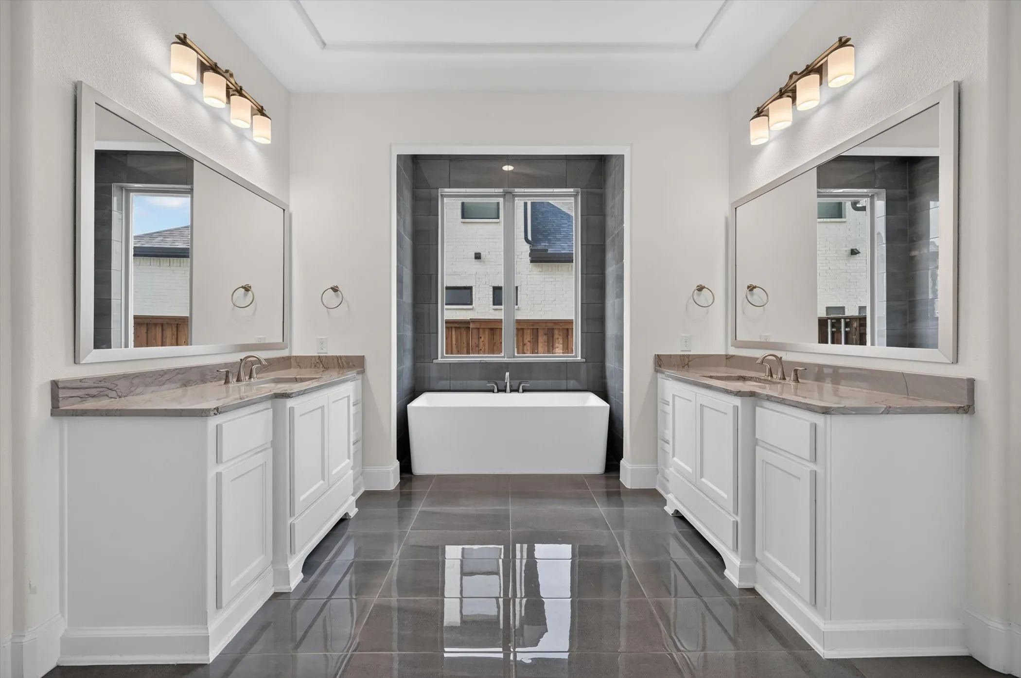 Spacious bathroom featuring a freestanding tub, dual vanities with white cabinetry, stone countertops, and large format floor tiling