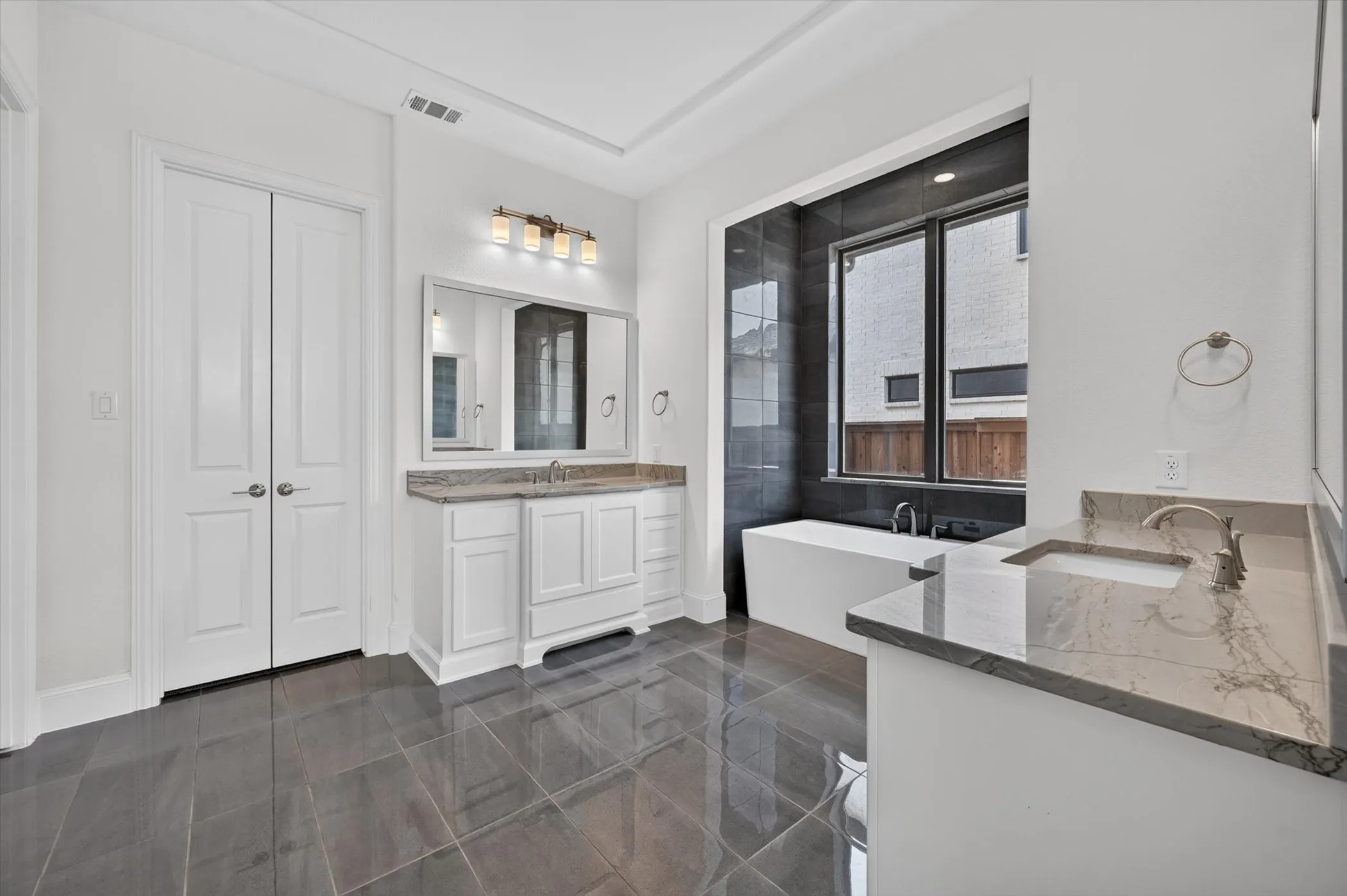Modern bathroom featuring a freestanding tub, dual vanities with stone countertops, dark tile flooring, and a large window with dark tile surround