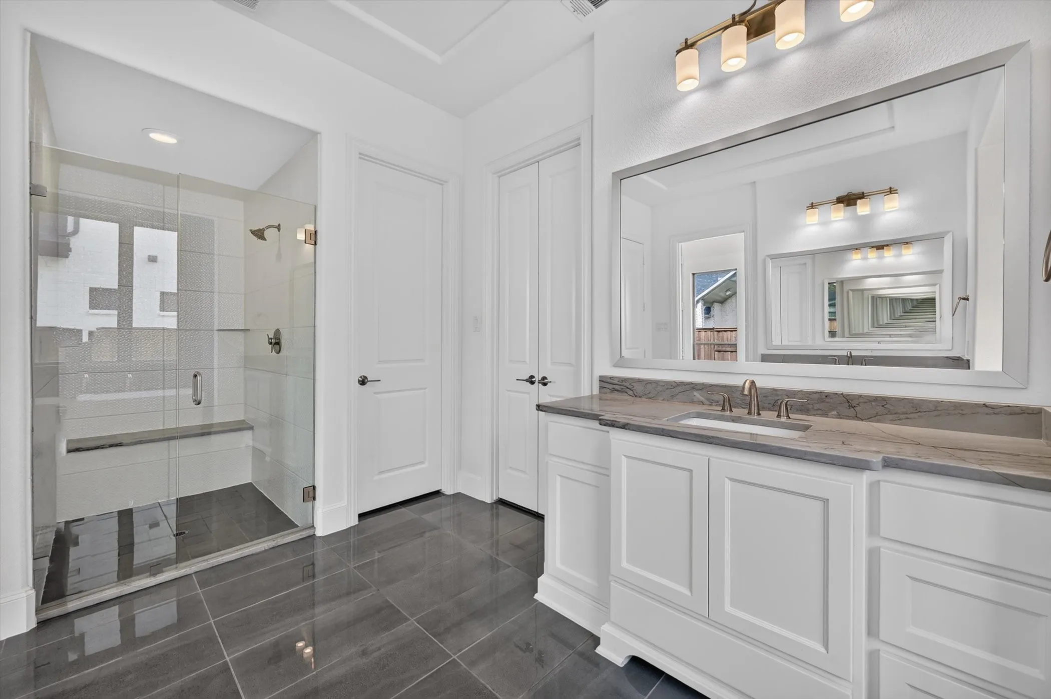 Spacious bathroom featuring a frameless glass shower enclosure with a built-in bench, a single vanity with a stone countertop and undermount sink, a large wall-mounted mirror, and dark gray tile flooring