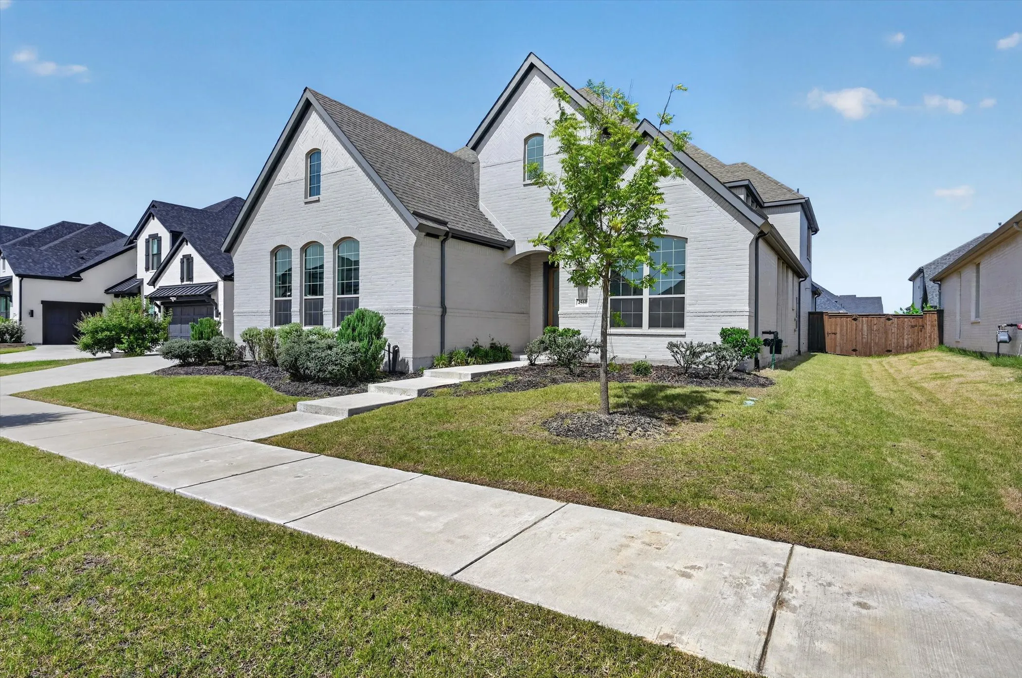 Contemporary-style residence featuring a light-colored brick exterior, dark gabled roofline, multiple arched windows, and a manicured front lawn with established landscaping
