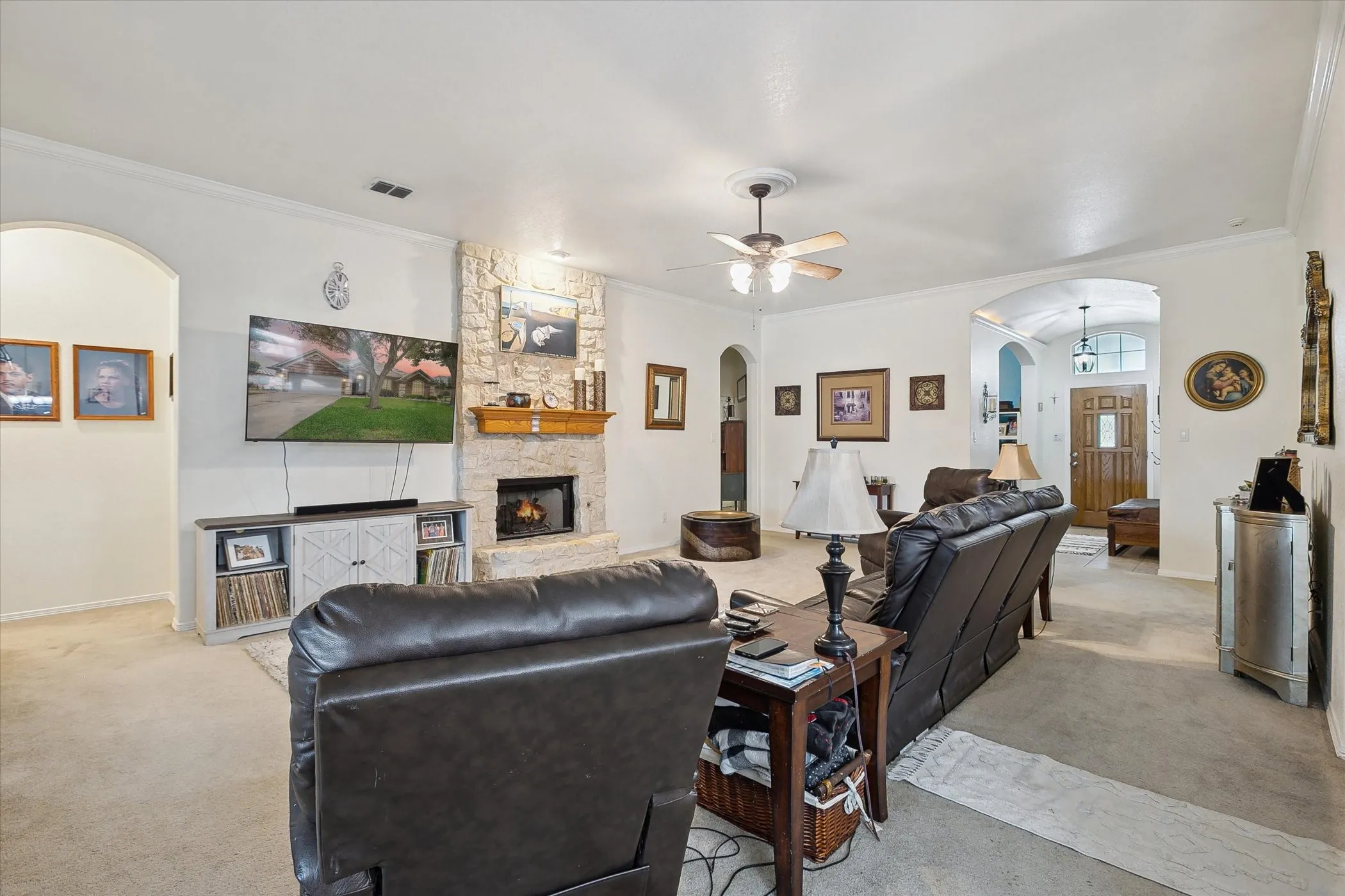 Spacious living room featuring a stone fireplace with a wood mantle, crown molding, and a ceiling fan