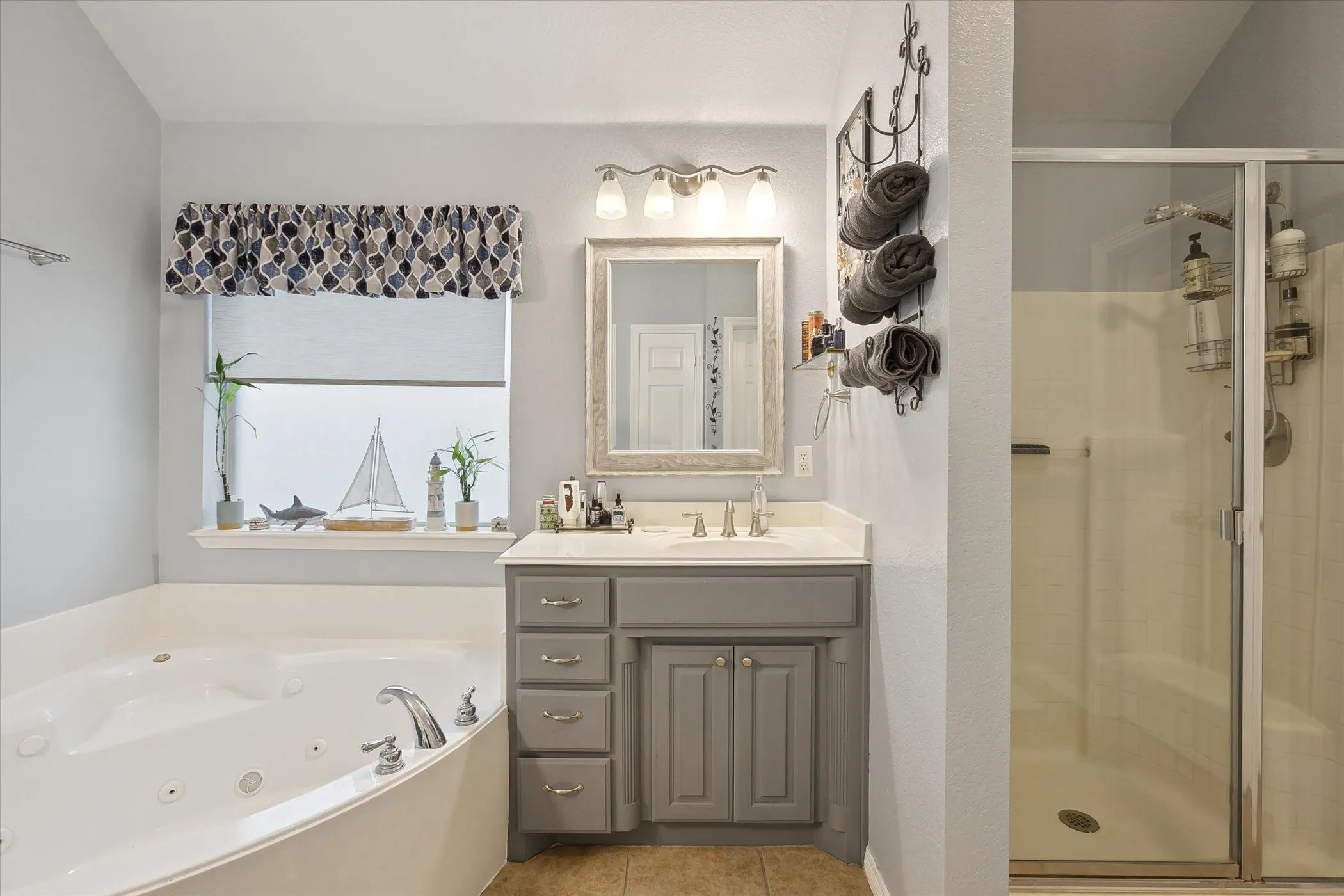 Primary bathroom featuring a corner jetted soaking tub, a vanity with a white countertop, a mirror with a decorative frame, a four-light vanity fixture, and a glass-enclosed shower