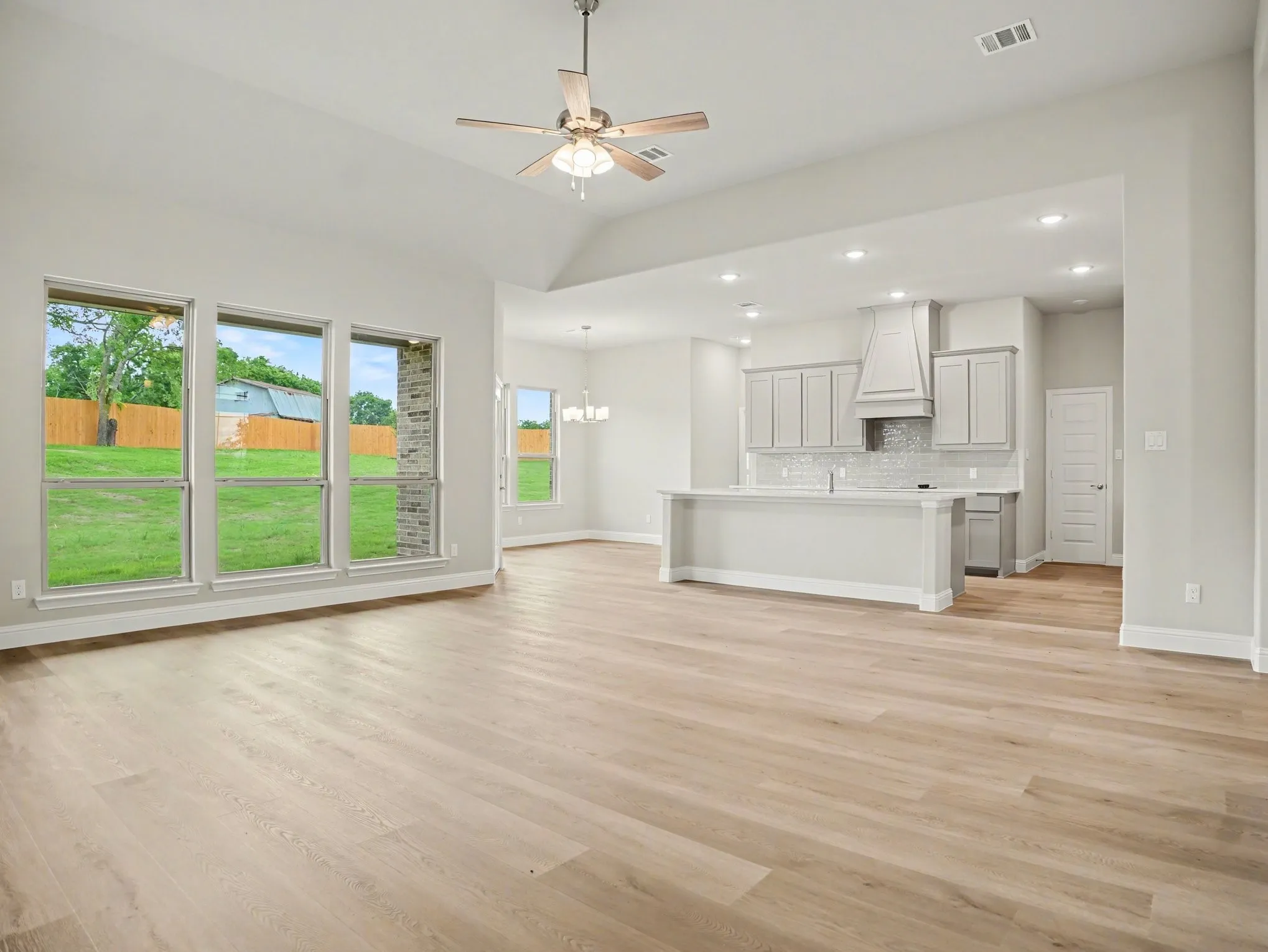 Open-concept living area featuring light wood-finish flooring, a vaulted ceiling with a ceiling fan, and multiple large windows