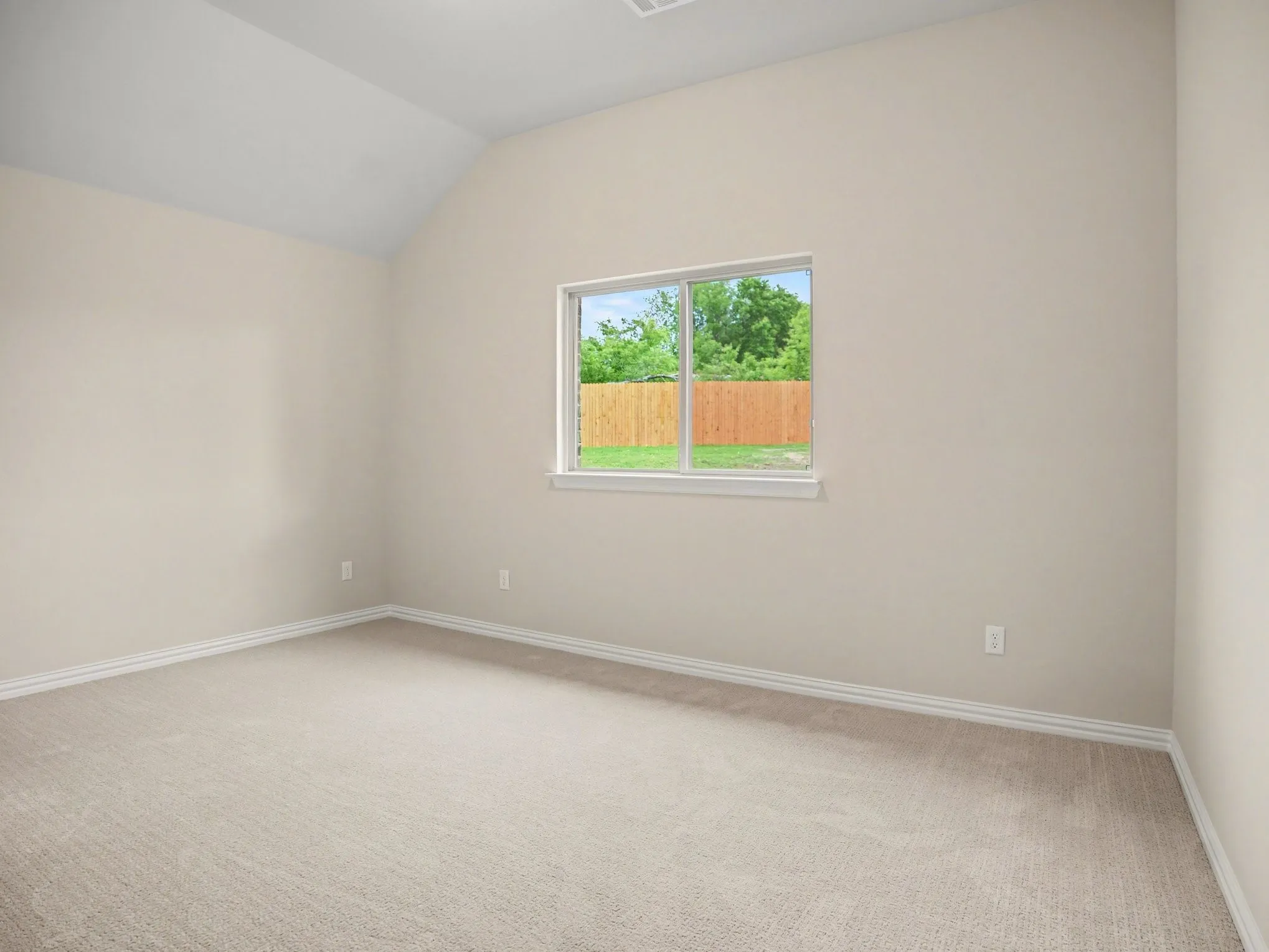 Vaulted ceiling bedroom featuring plush neutral carpeting, white baseboards, and a large window with a wood fence backdrop