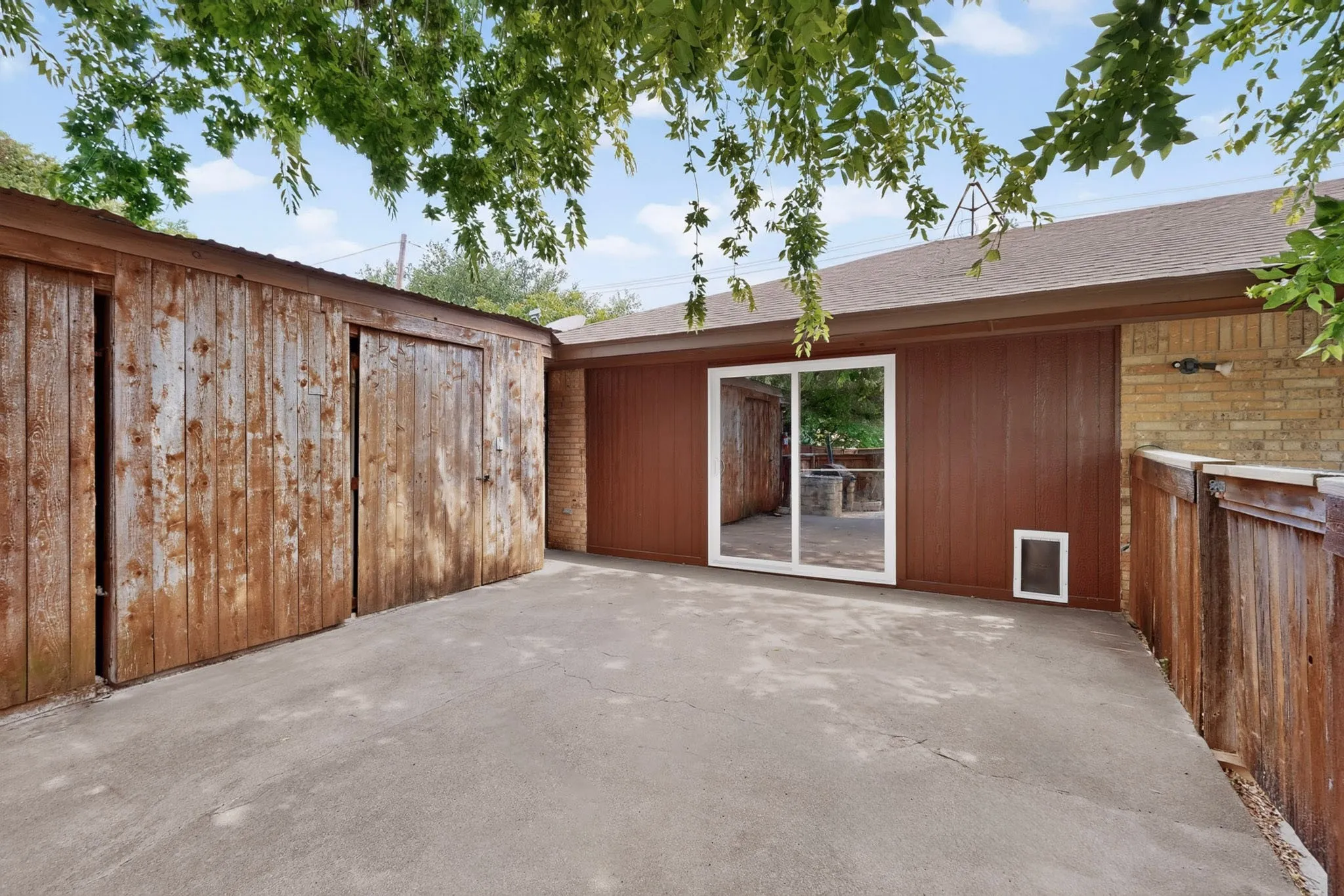 Concrete patio area featuring a shed with sliding wooden doors, a solid brown exterior wall, and a sliding glass door