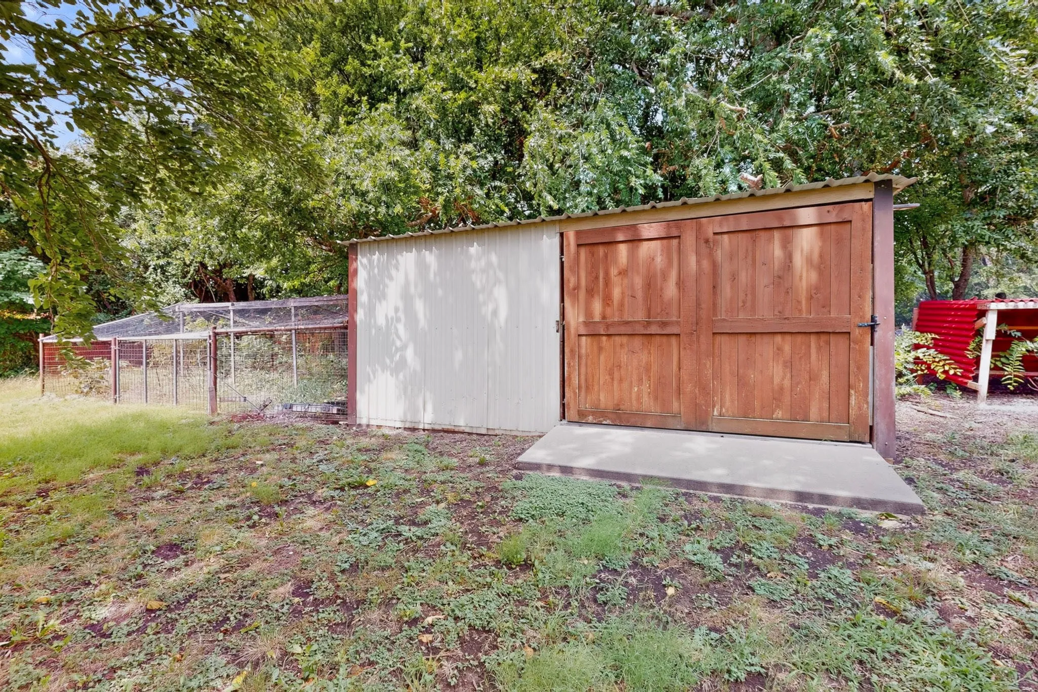 Freestanding shed with wood double doors, a concrete pad, and a metal roof, accompanied by an enclosed structure with wire fencing