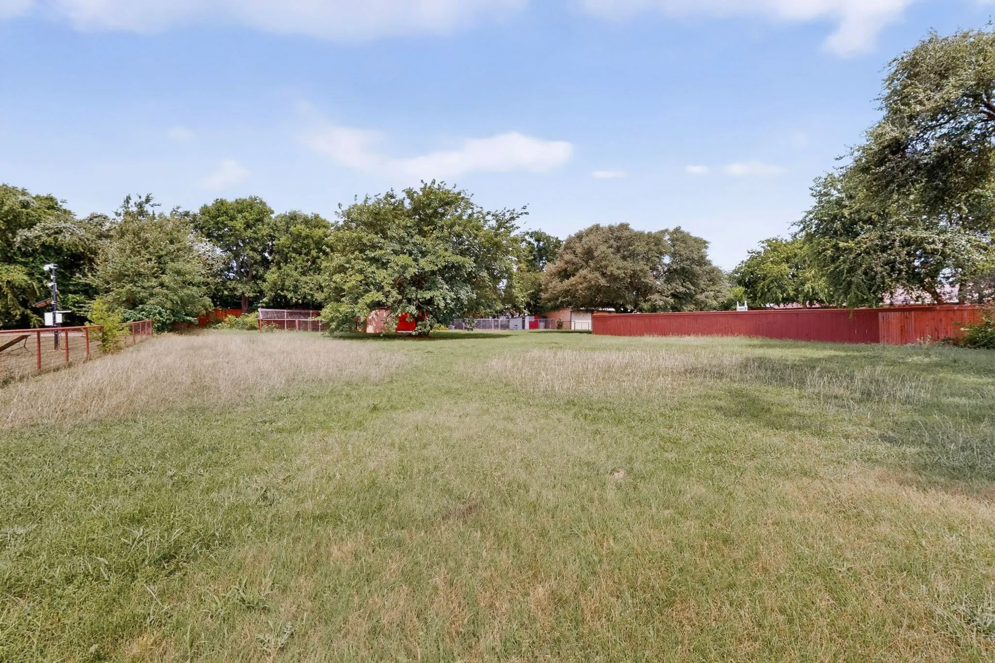 Expansive lot featuring a perimeter fence, mature trees, and a clear blue sky overhead