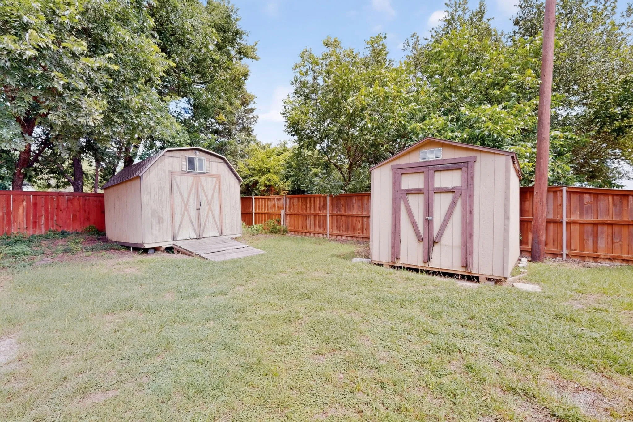 Expansive backyard featuring two utility sheds, a brown privacy fence, and mature trees
