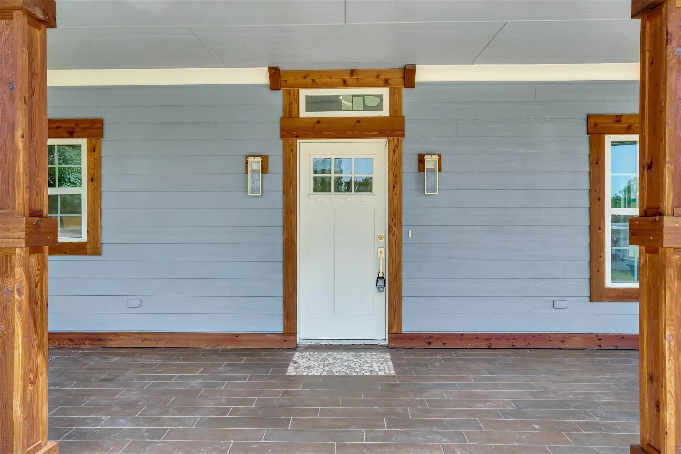 Inviting front porch featuring wood-finish flooring, light gray siding, white entry door with transom window, and natural wood trim accents