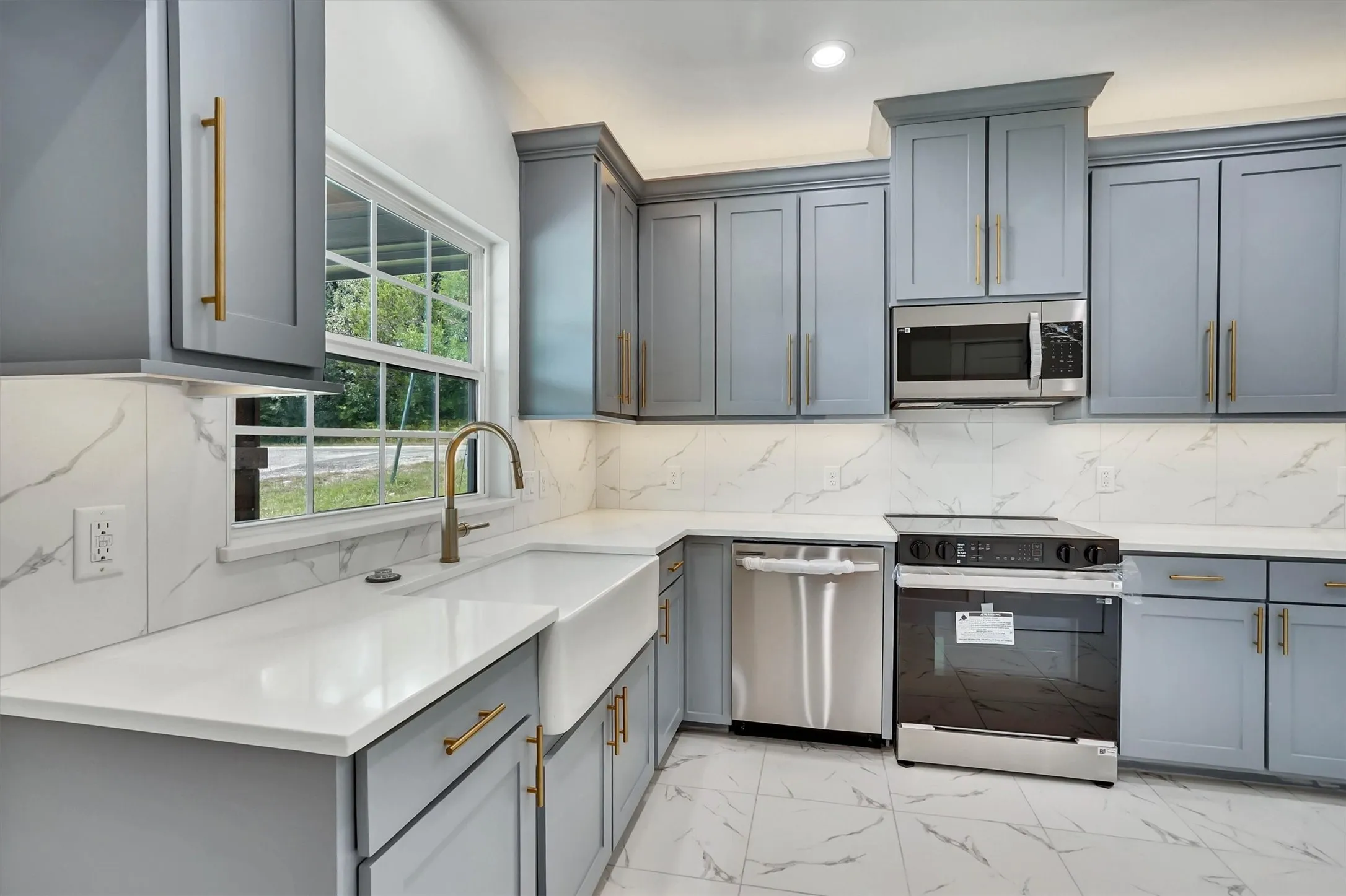 Modern kitchen featuring light gray blue cabinetry, gold-tone hardware, quartz white countertops, a white farmhouse sink, and a stainless steel appliance package