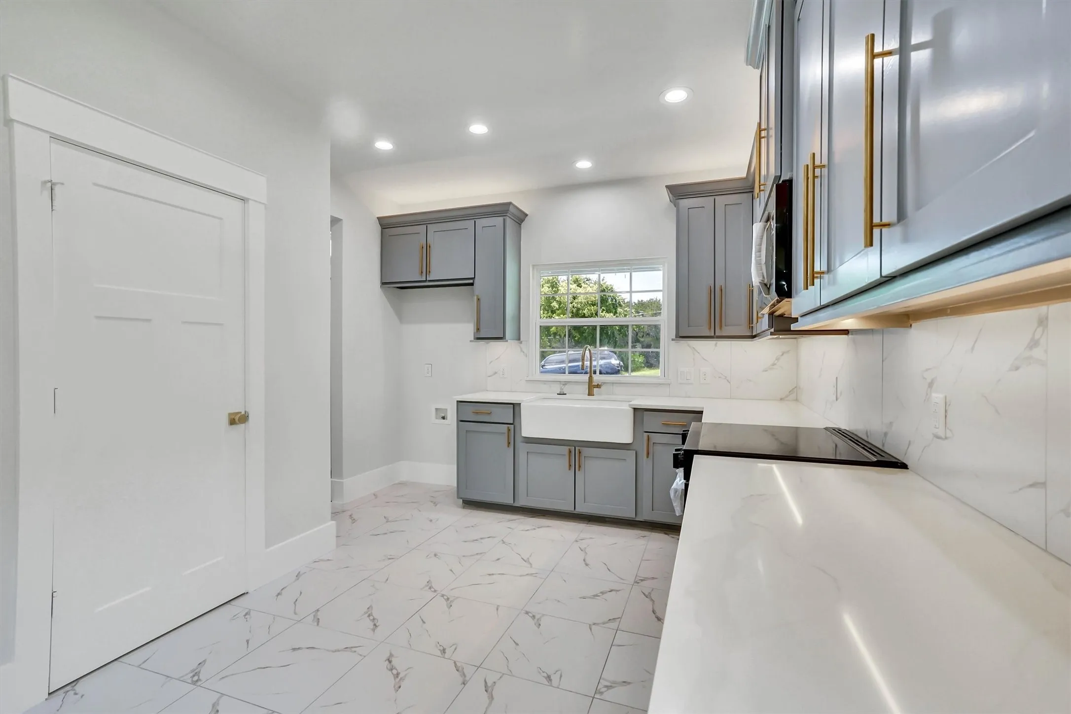 Kitchen featuring light grey cabinetry with gold-finish hardware, a white farmhouse sink with a gold-finish faucet, and a black cooktop