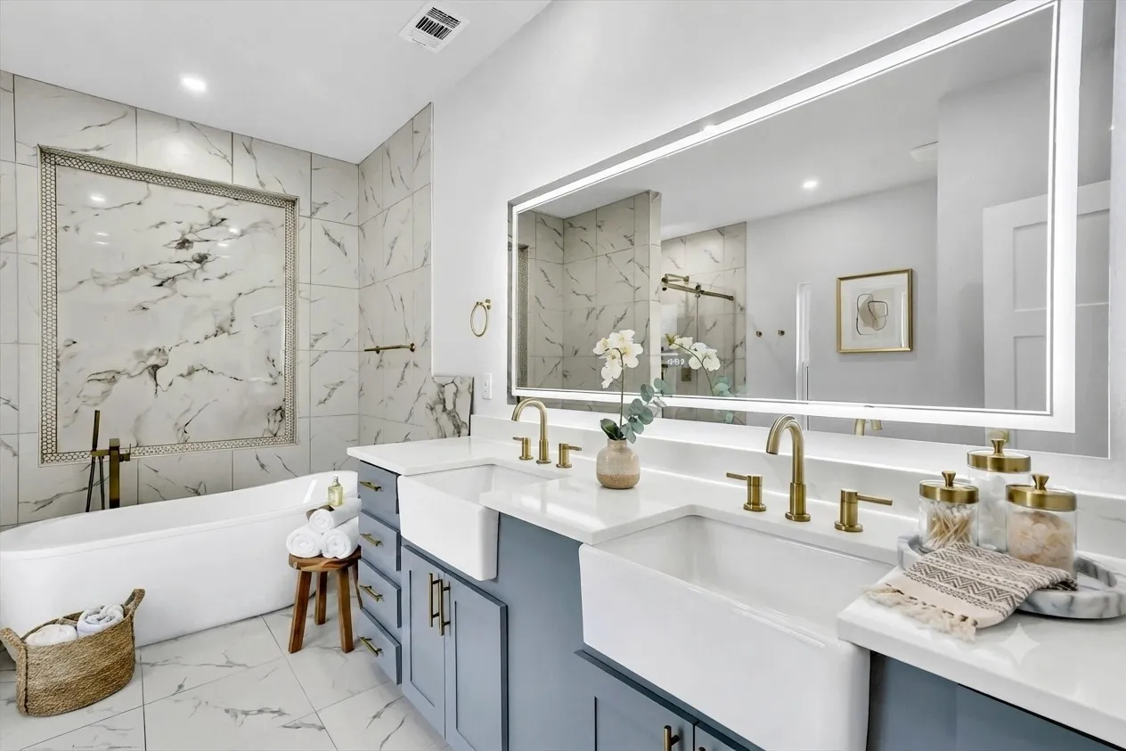 Contemporary bathroom featuring a freestanding tub, dual farmhouse sinks, light gray-blue vanity cabinetry, and a large backlit mirror