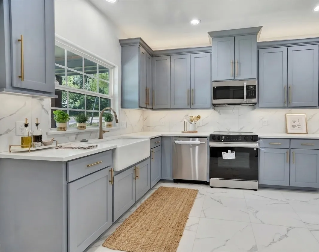 Modern kitchen featuring light gray cabinetry with gold hardware, white countertops, a deep farmhouse sink, stainless steel appliances, and white marble-look tile flooring