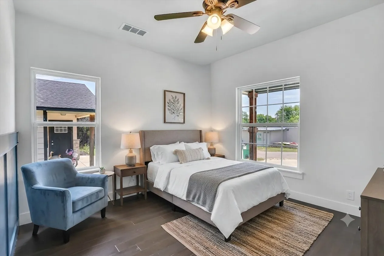 Bedroom featuring dark wood-finish flooring, two large windows, a ceiling fan with integrated lighting, and white walls