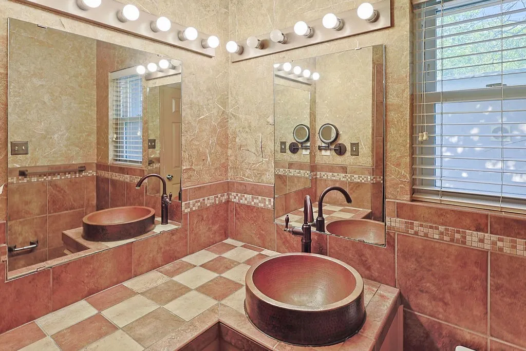 Bathroom featuring dual vessel sinks with bronze-finish fixtures, a window with blinds, full-height wall tiling, and a tiled vanity surface
