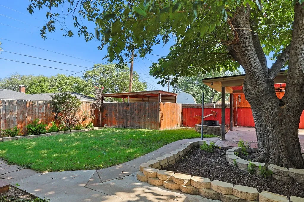 Spacious backyard featuring a covered patio, mature tree, stone-lined planter beds, and a perimeter wood fence