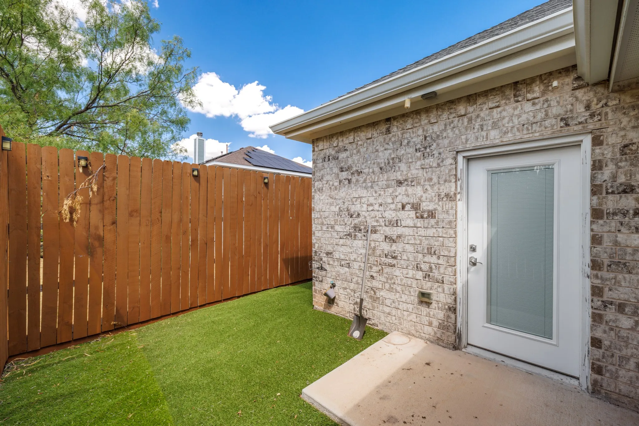 Low-maintenance synthetic turf, a wood privacy fence with lighting, and a brick exterior with a rear entry door