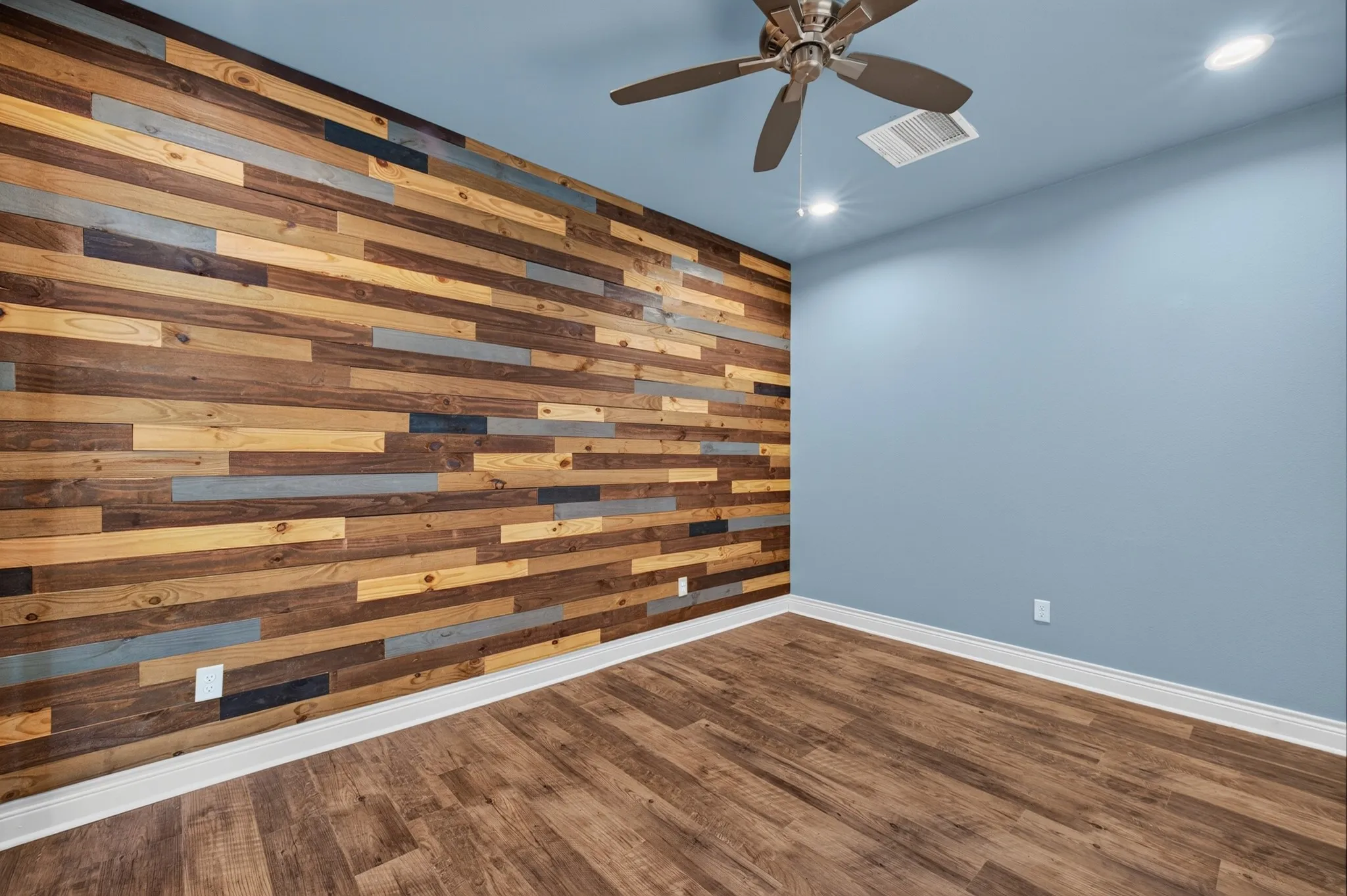 Wood-finish flooring, accent wall with varied wood paneling, ceiling fan with integrated lighting, recessed ceiling lights, pale blue wall finish