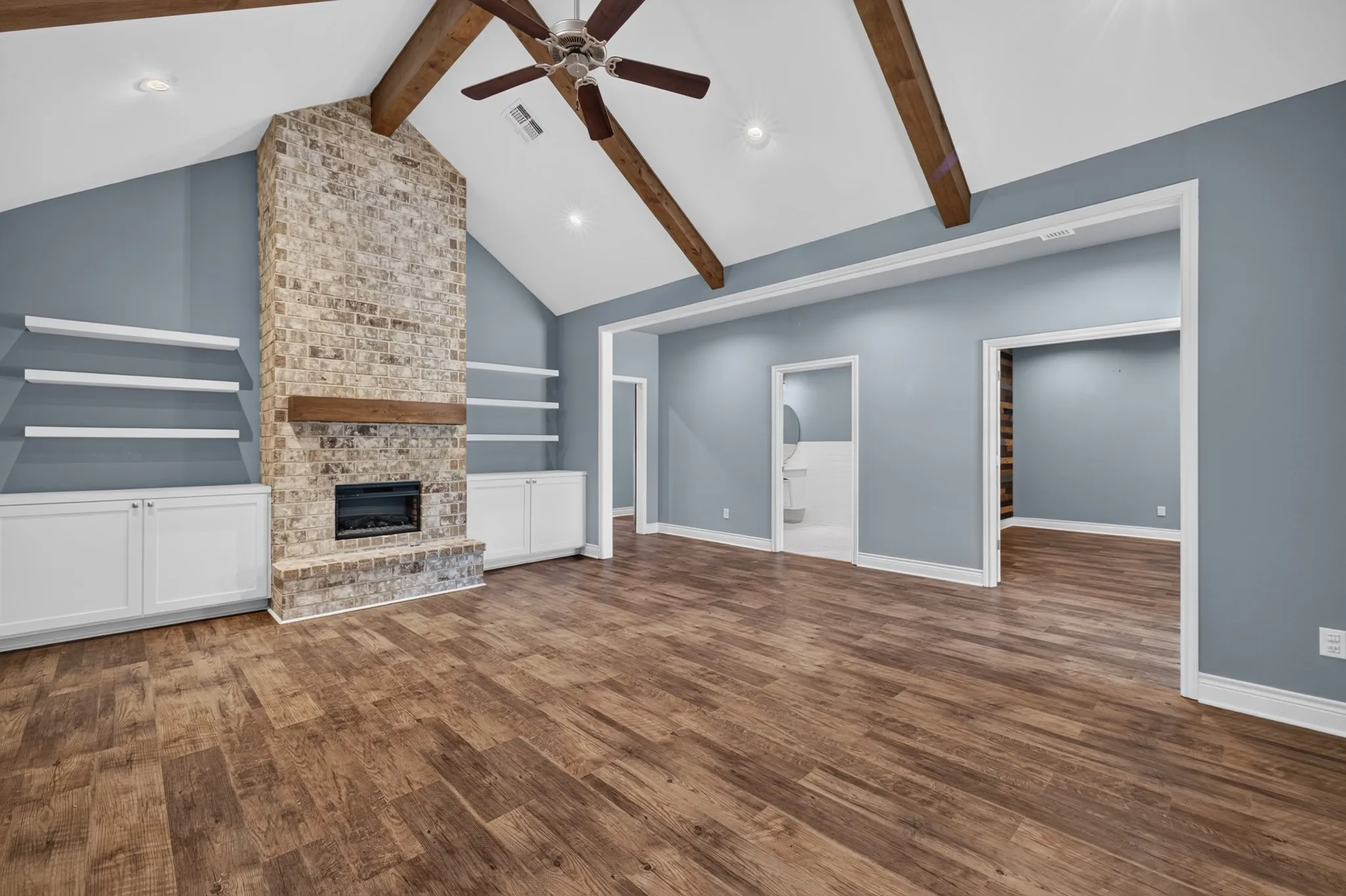 Vaulted ceiling with exposed wood beams, wood-finish flooring, recessed lighting, brick fireplace with wood mantel, and built-in cabinetry with shelving