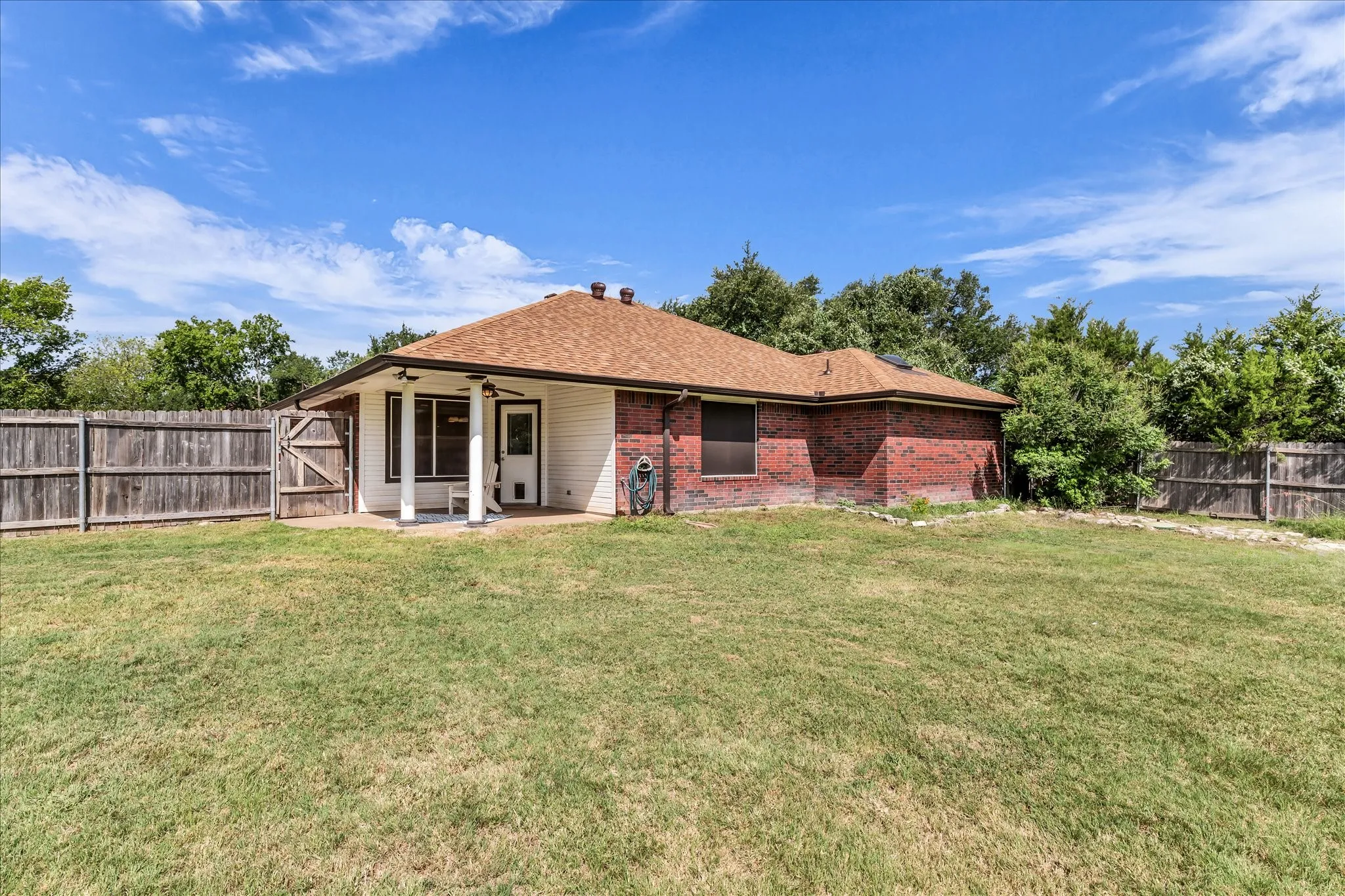 Expansive backyard featuring a covered patio, brick exterior, wood fence, and mature perimeter trees