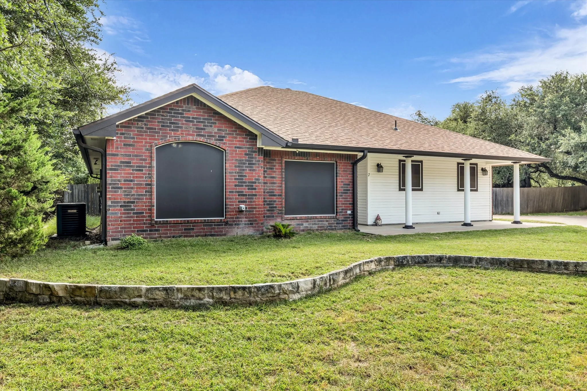 Brick and white siding exterior featuring a covered patio with white columns, a stone retaining wall, and a shingled roof