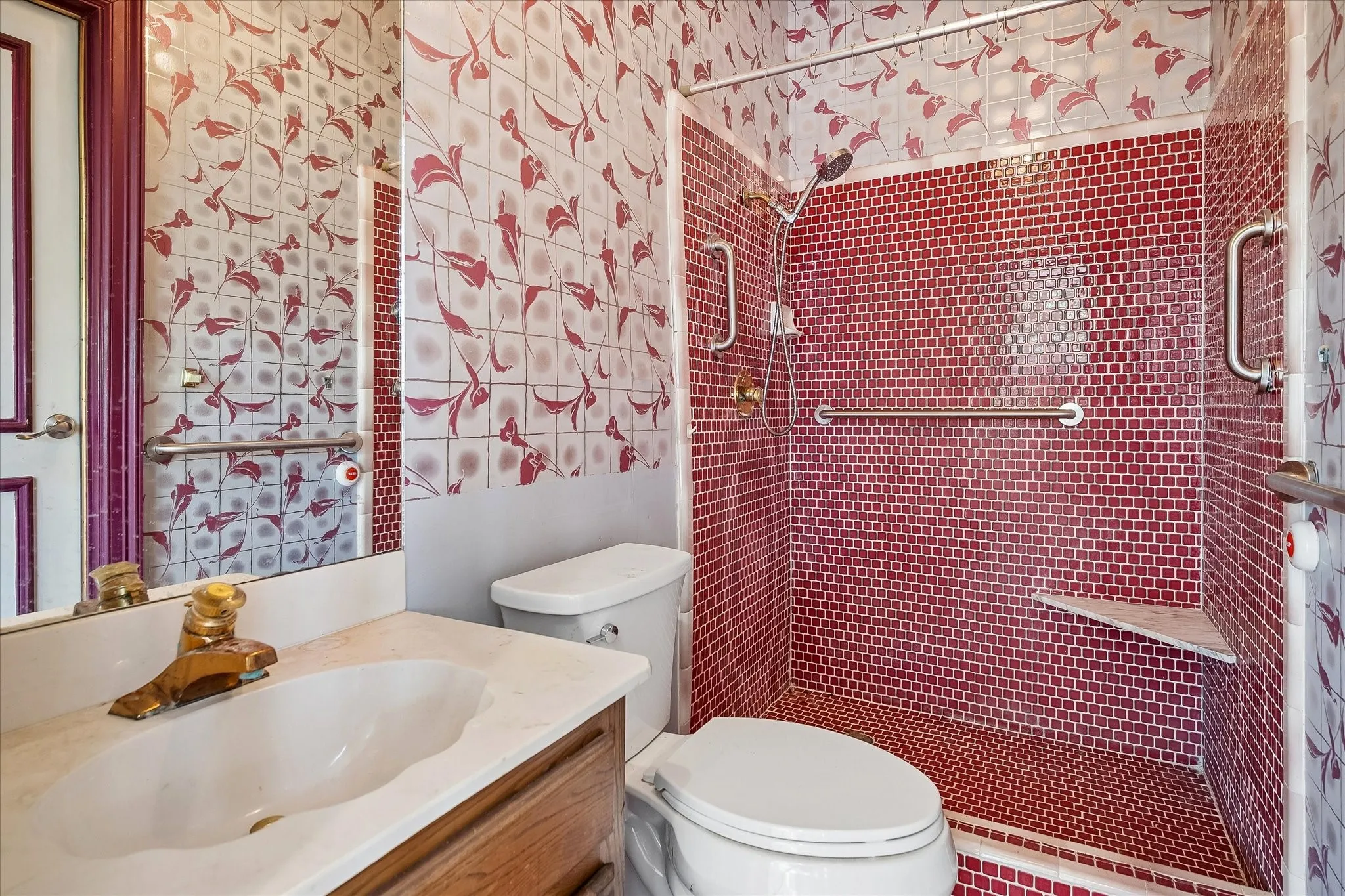 Full bathroom featuring a walk-in shower with red mosaic tile and grab bars, a built-in shower bench, and a vanity with a white basin and brass-finish faucet