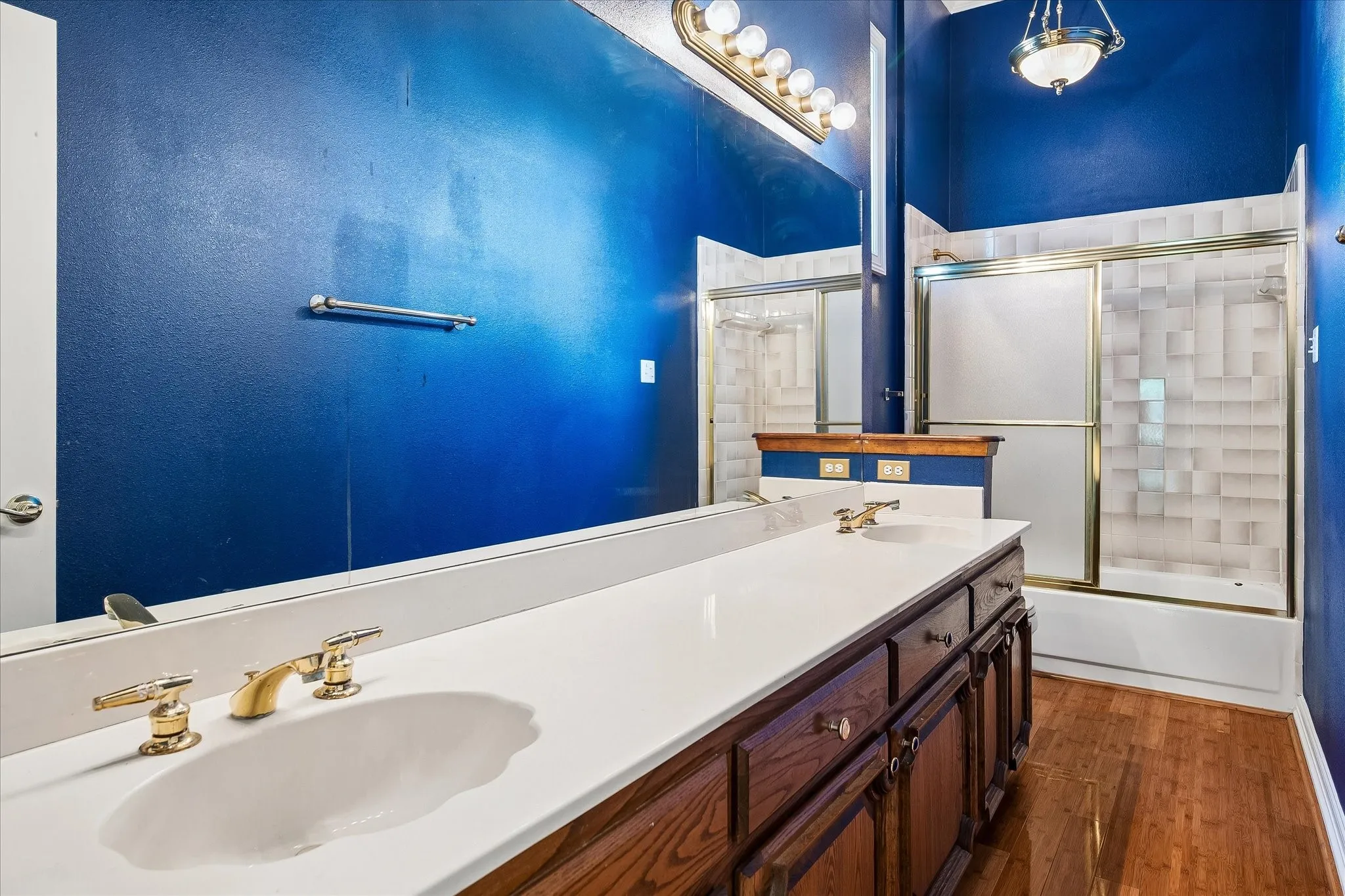 Double vanity with white countertop, brass-finish faucets, wood-finish cabinetry, and full-width mirror