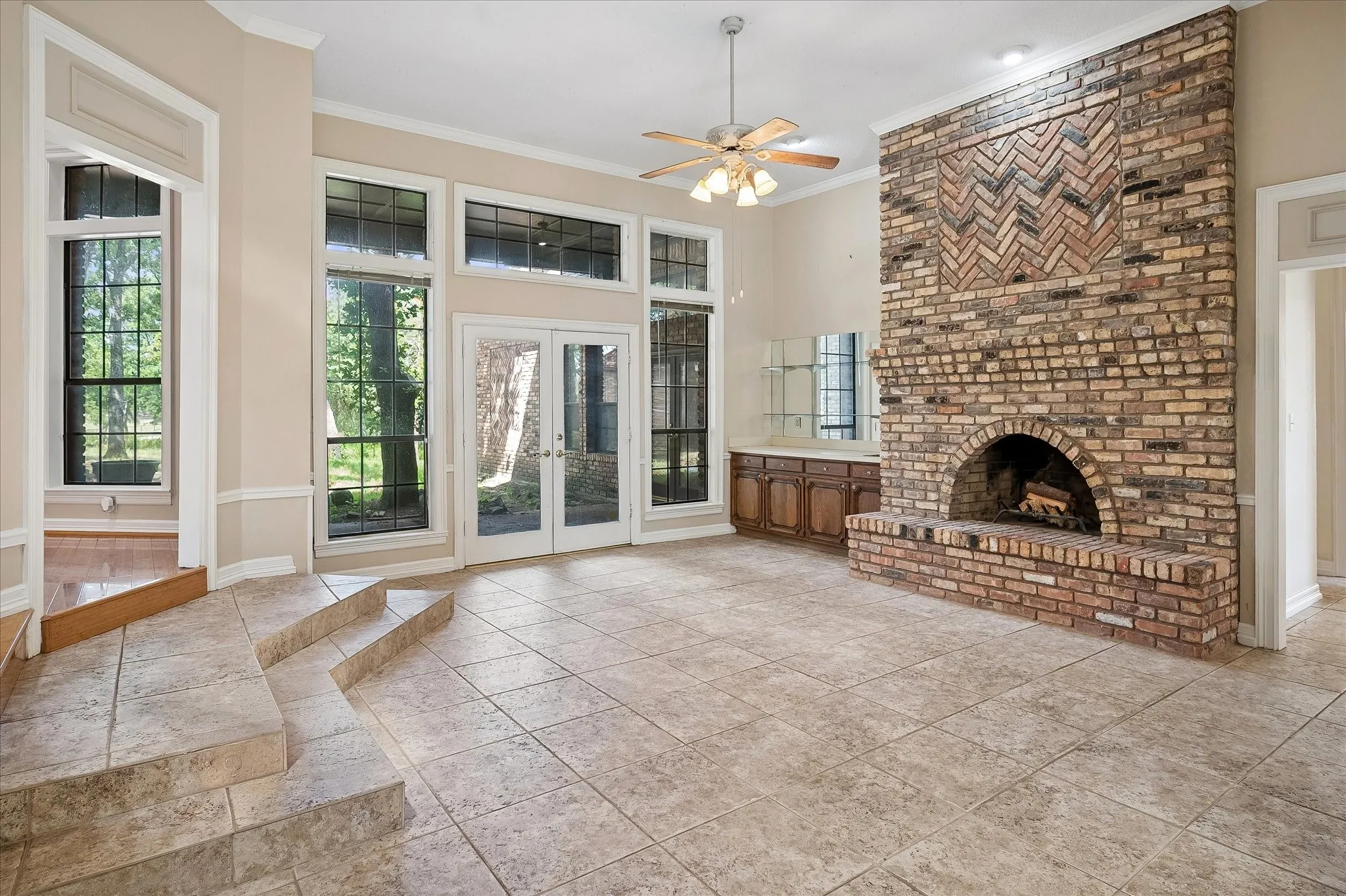 Spacious living area featuring a floor-to-ceiling brick fireplace with a decorative herringbone pattern, multiple large windows, glass-paned double doors, tiled flooring, and a built-in wet bar with cabinetry