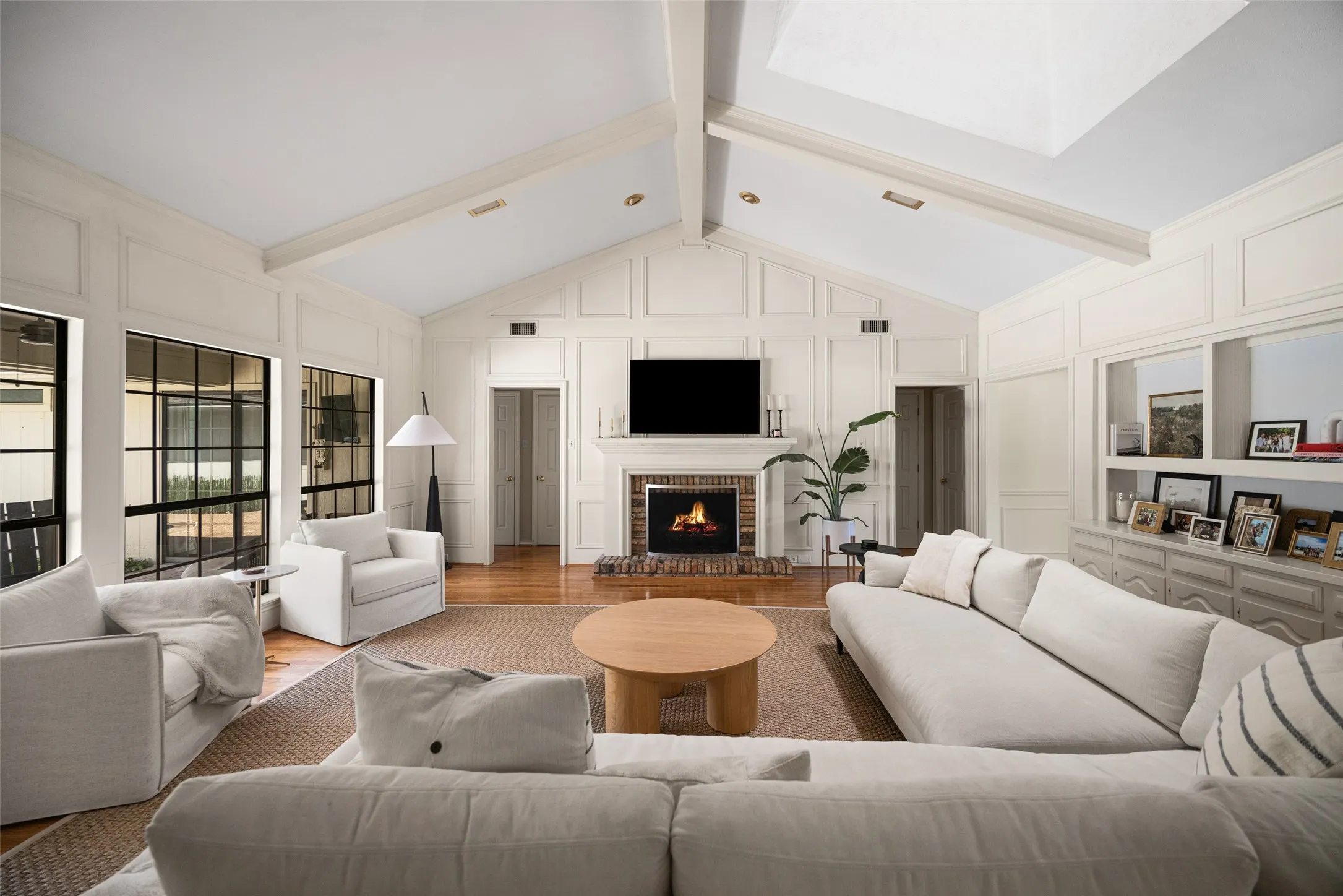 Living area featuring a brick fireplace with a mantle, vaulted ceilings with exposed beams, wood-finish flooring, and large grid-pattern windows