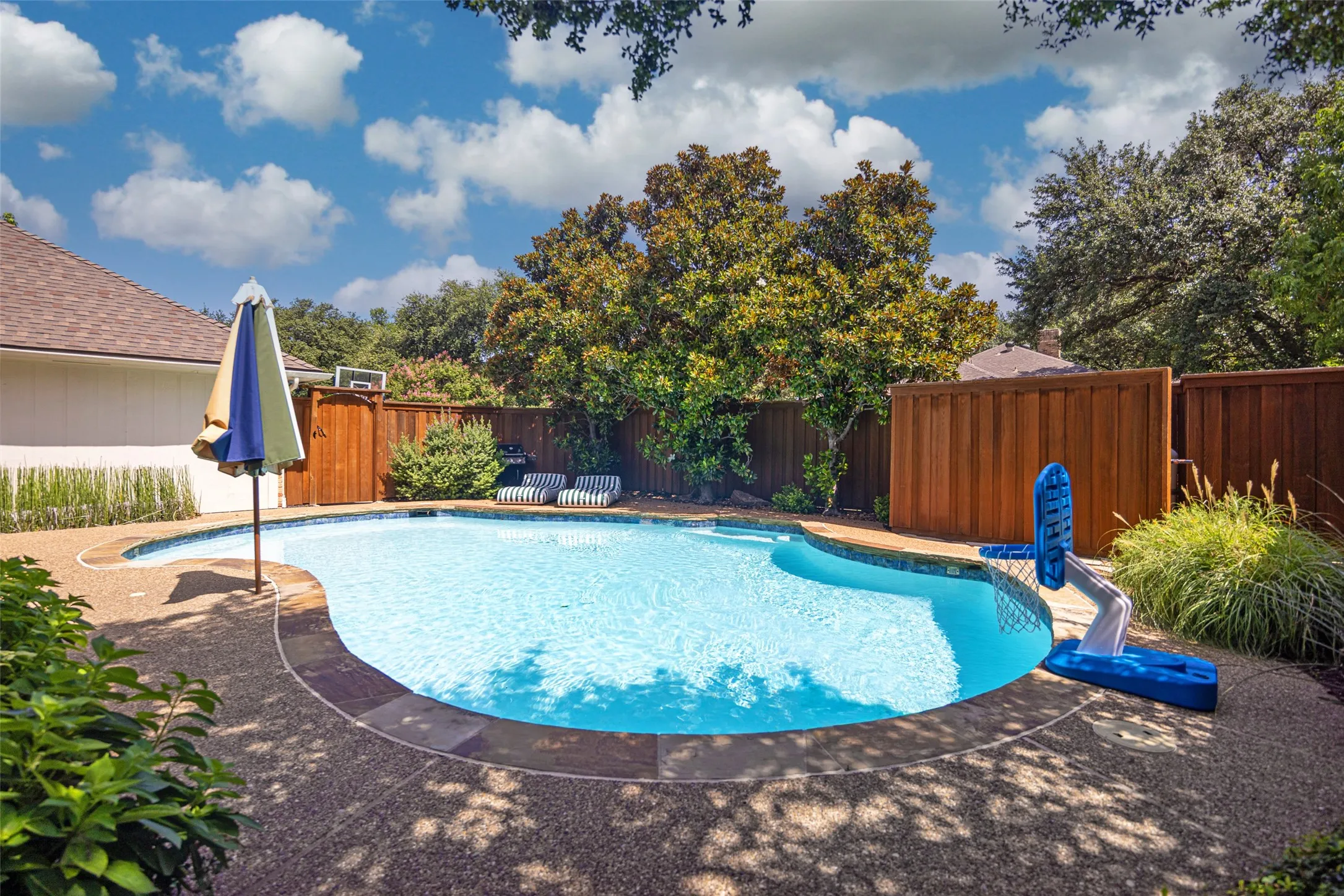Curved swimming pool with an exposed aggregate deck, bordered by mature trees and privacy fencing