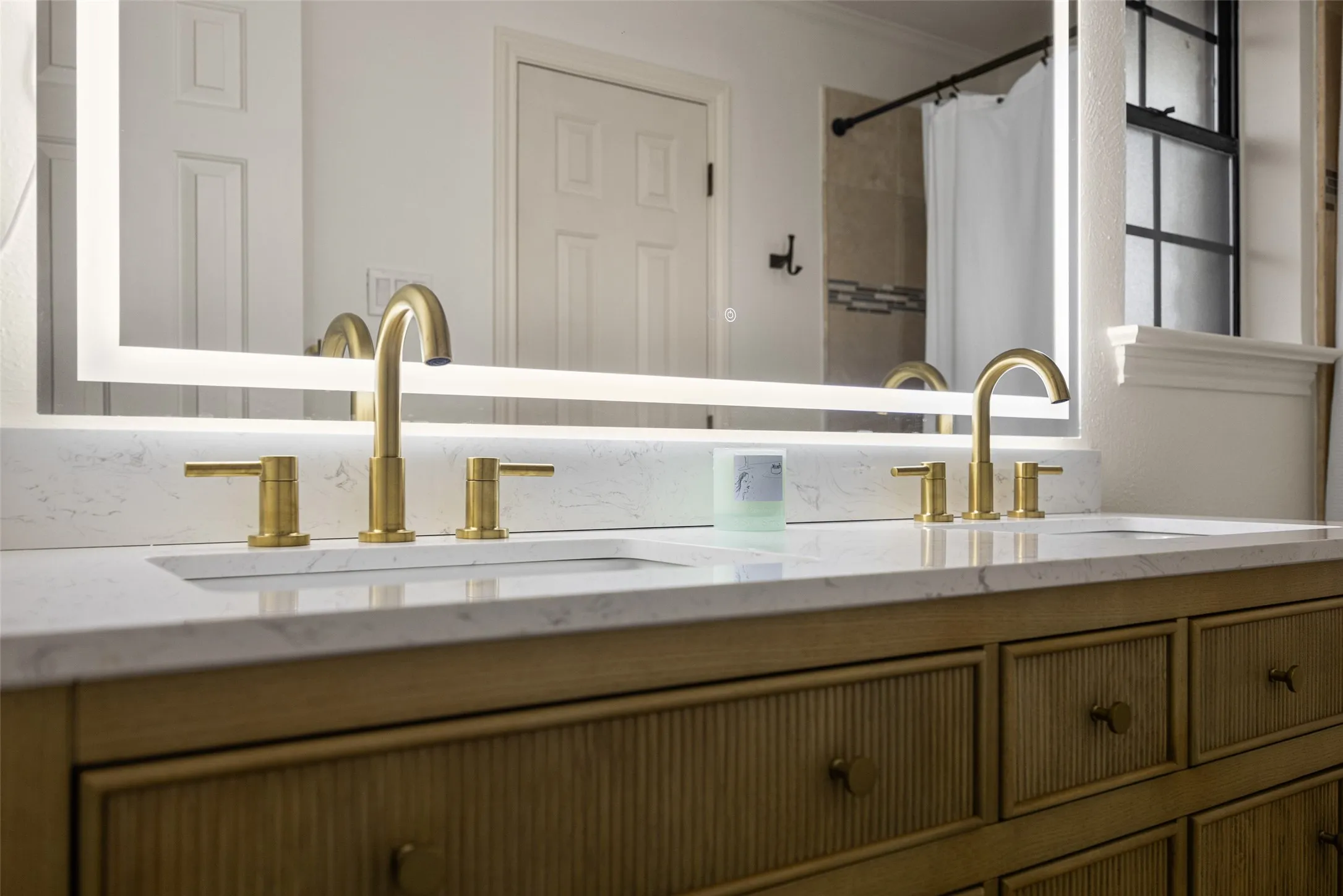 Dual vanity featuring a white countertop with integrated sinks, brushed gold faucets, and a large backlit mirror