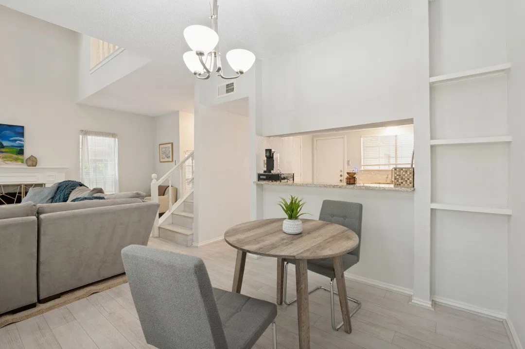 Open-concept dining and living area featuring a round wood-finish table, gray upholstered chairs, light wood flooring throughout, and a modern pendant light fixture