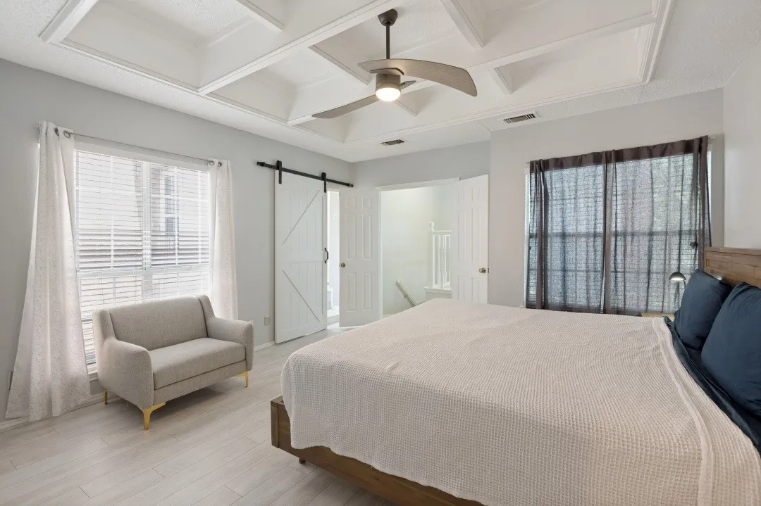 Primary Bedroom Suite with coffered ceiling, ceiling fan with brushed metal finish, wood-finish flooring throughout, and multiple windows with operable shutters and sheer curtains