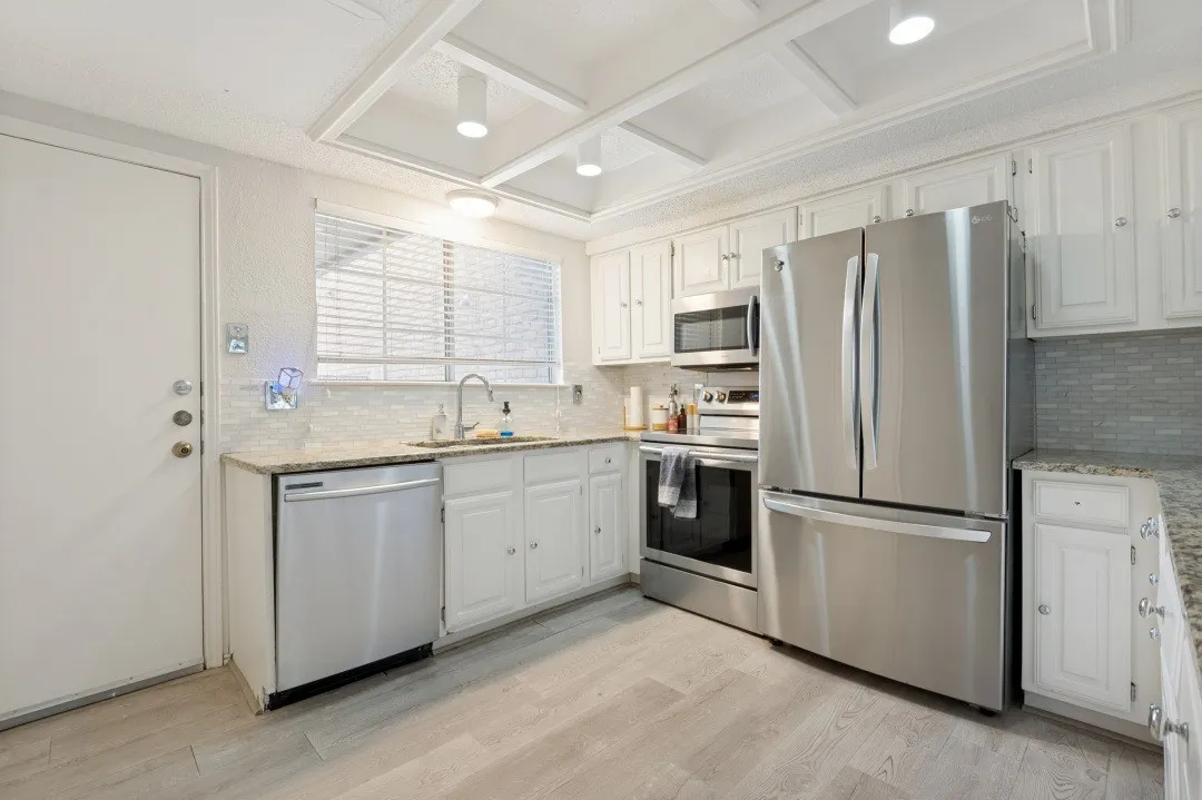 Modern kitchen featuring stainless steel appliances, white cabinetry with ample storage, wood-finish flooring, granite countertops, tile backsplash, recessed ceiling lighting, and a functional layout with sink, cooktop, and built-in microwave