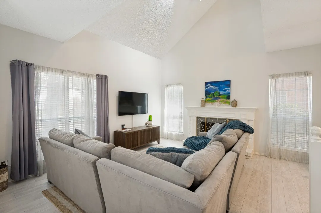 Bright, open-concept living room showcasing vaulted ceilings, abundant natural light from multiple windows with sheer and panel drapes, light wood-finish flooring, and a built-in fireplace with shelving