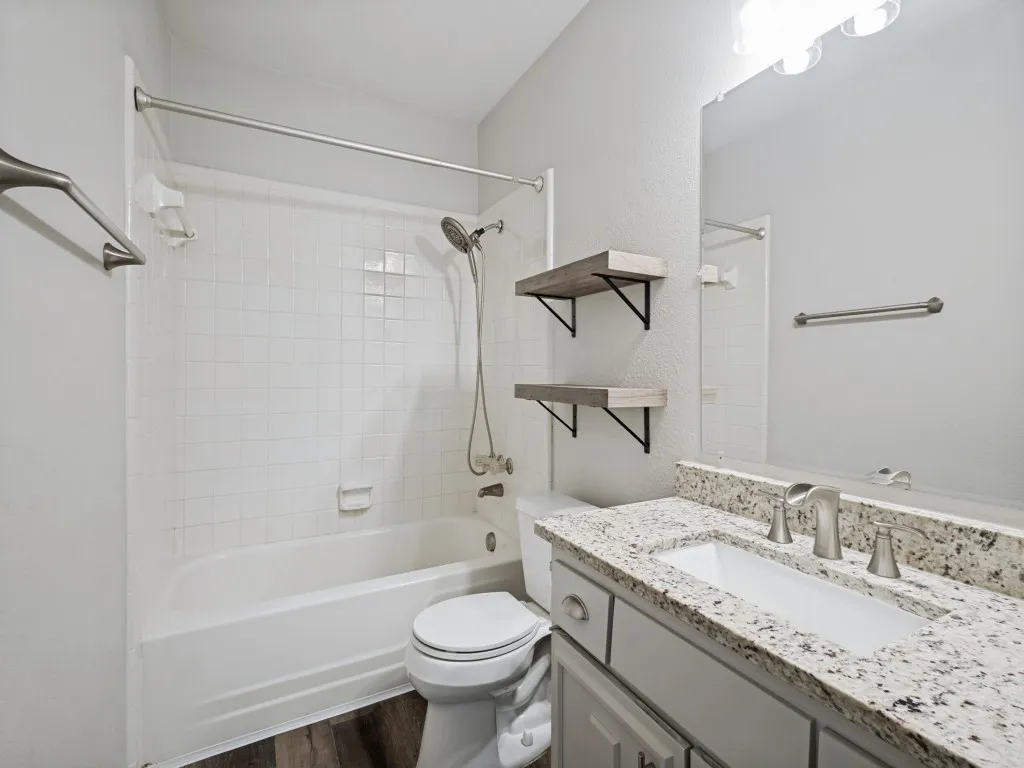 Full bathroom featuring a white tub with subway tile surround, brushed nickel fixtures, a vanity with a stone countertop and undermount sink, and wood-finish flooring