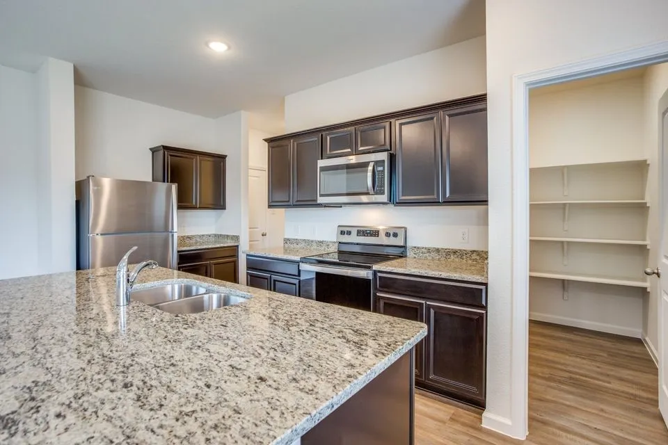 Kitchen featuring an island with a stainless steel double basin sink, dark wood-finish cabinetry, stainless steel appliances, speckled stone countertops, and wood-finish flooring
