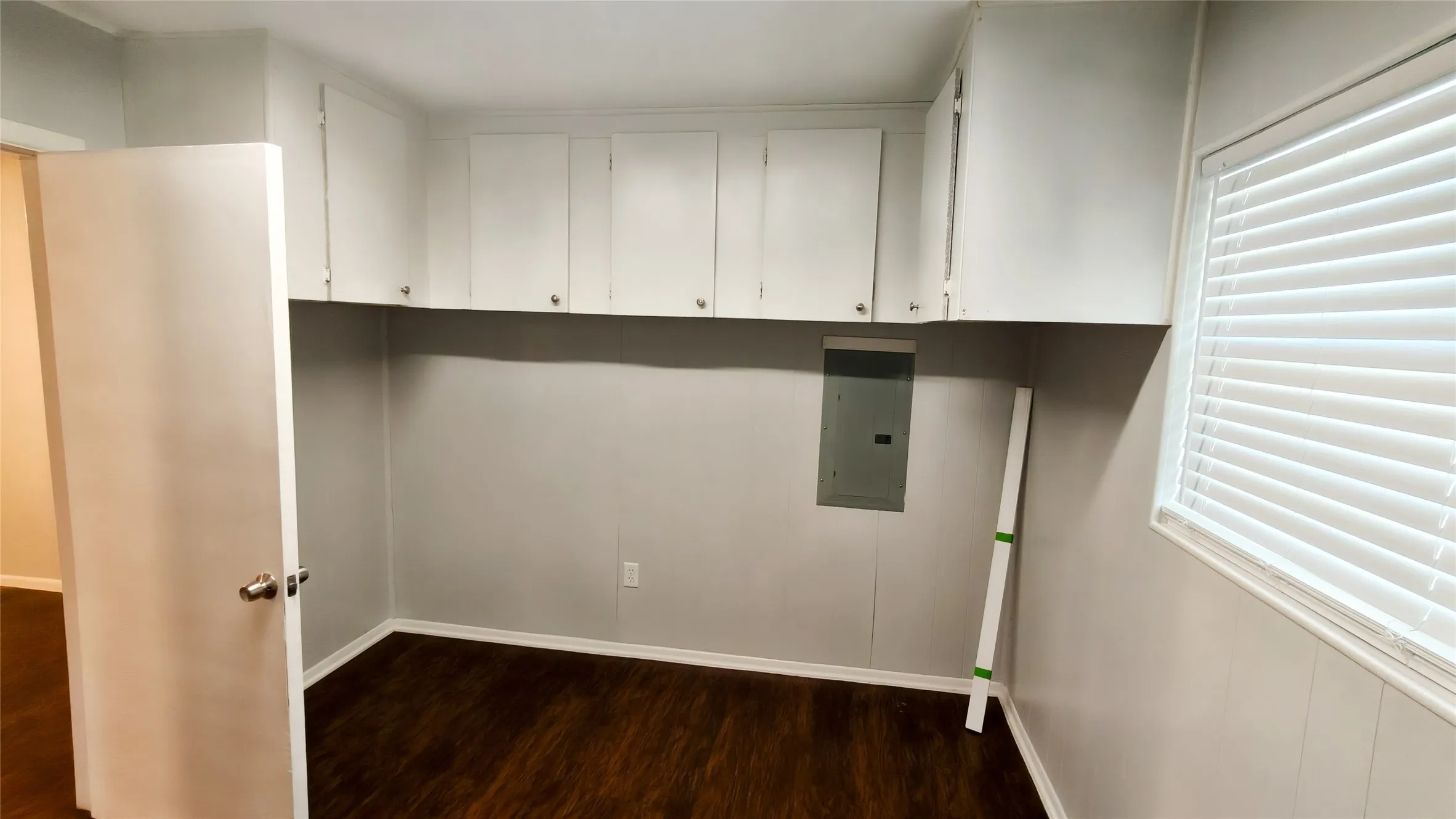 Room with overhead white cabinetry, wood-finish flooring, a white door, and a window with blinds