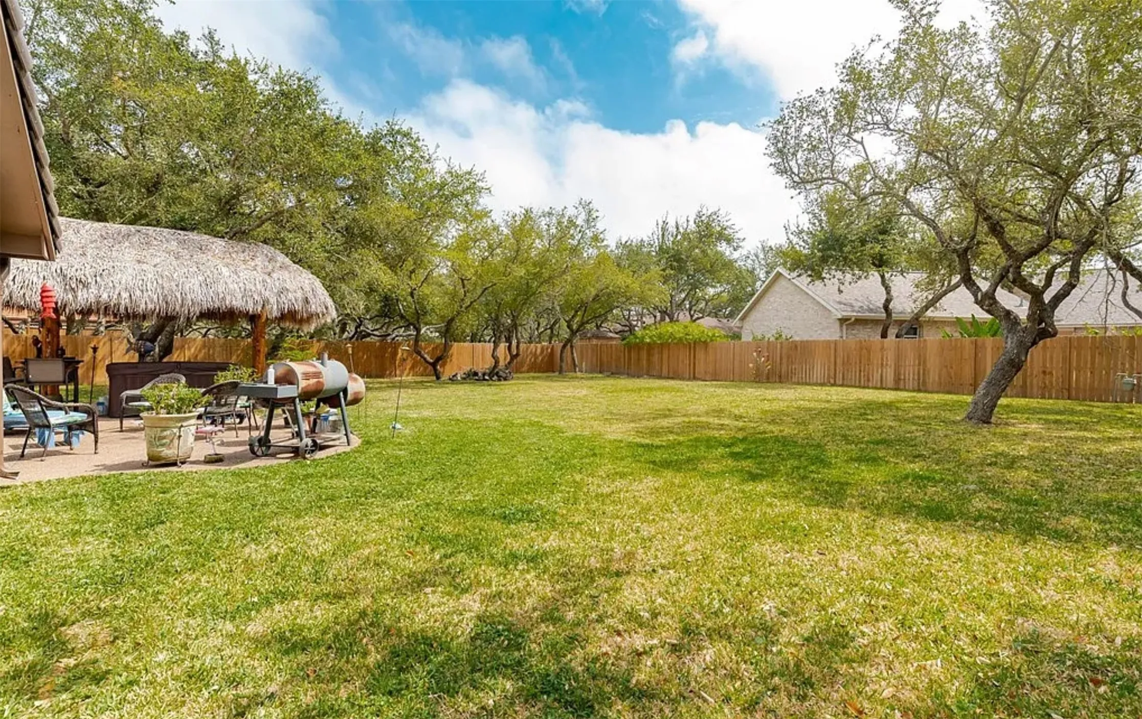 Expansive backyard featuring a large grass area, mature trees, a thatched-roof palapa, and a privacy fence