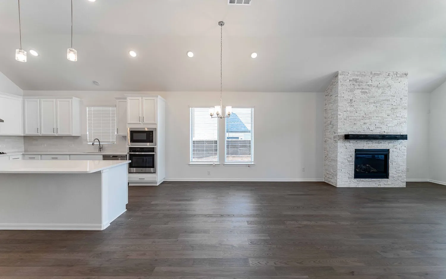 Open-concept living space featuring wood-finish flooring, a contemporary fireplace with stacked stone surround and dark mantel, and a kitchen with white cabinetry, built-in appliances, and a central island