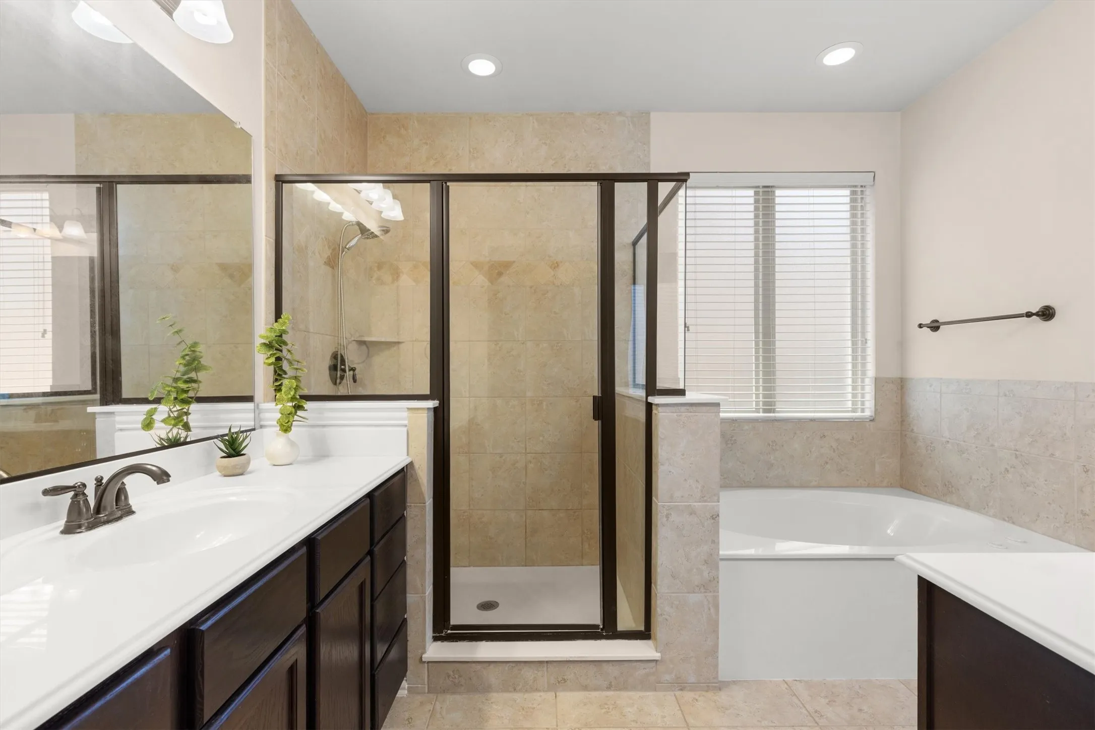 Bathroom featuring a glass-enclosed shower, a built-in tub with tile surround, a dark wood-finish vanity with dual sinks and white countertops, and a window with blinds