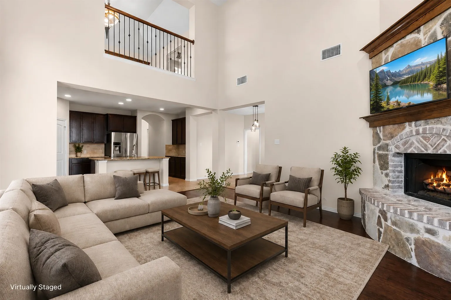 Soaring ceiling, stone fireplace with hearth, dark wood-finish flooring, open-concept kitchen with breakfast bar, and an upper-level balcony with metal balusters